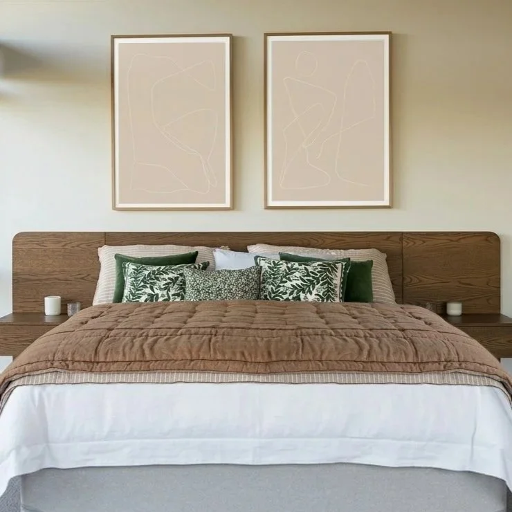 A bedroom with a bed featuring green and white patterned pillows, a brown quilt, and a white bed sheet. There are two wall art pieces with abstract line drawings hanging above the bed, and small candles or decorative items on either side of the headboard.