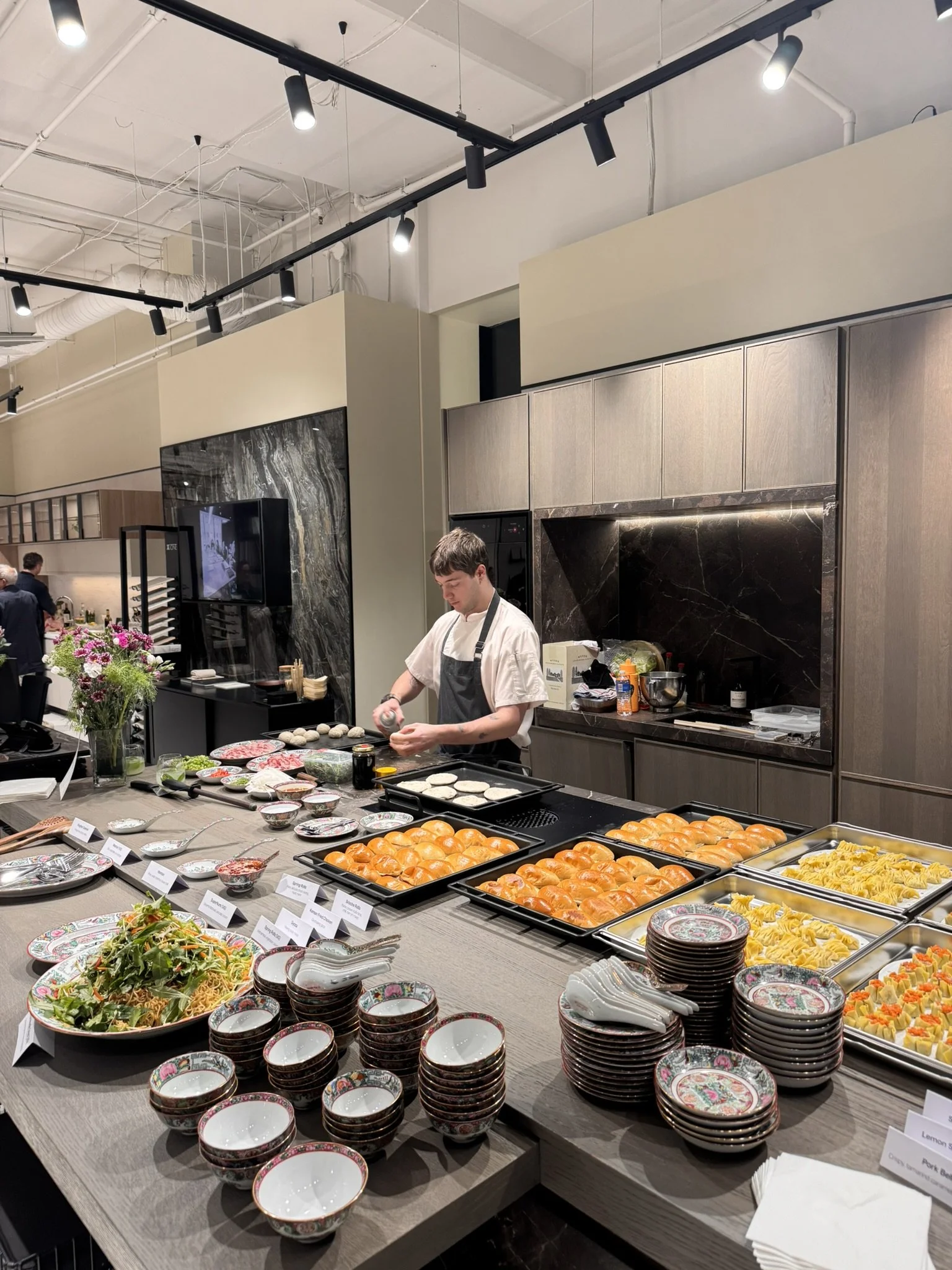 A chef prepares food at a buffet table filled with various dishes, including salads, pasta, bread rolls, and sliced meats, in a modern, well-lit restaurant or event space.