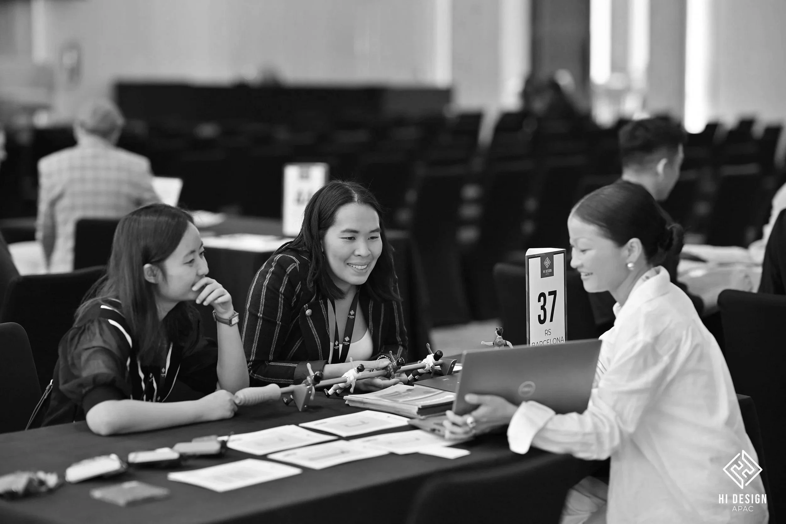 Three women sitting at a table, playing foosball during an event or convention, with a sign indicating table number 37.