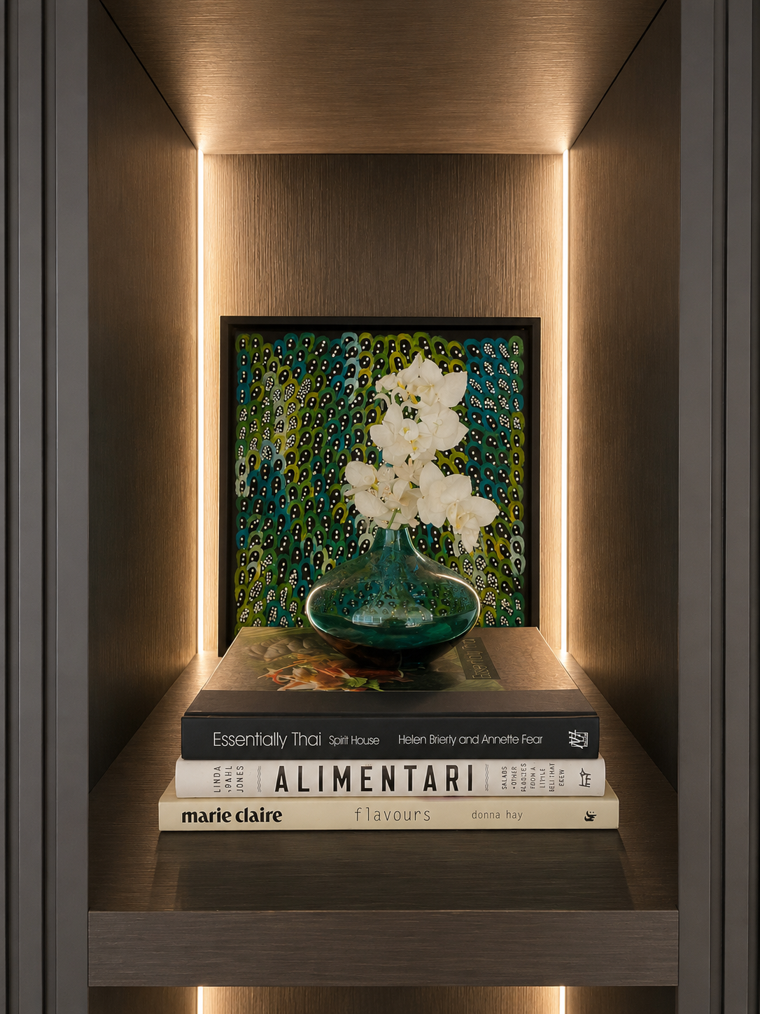 Decorative display shelf with a vase of white orchids, a stack of three cookbooks, and a patterned art piece in the background.