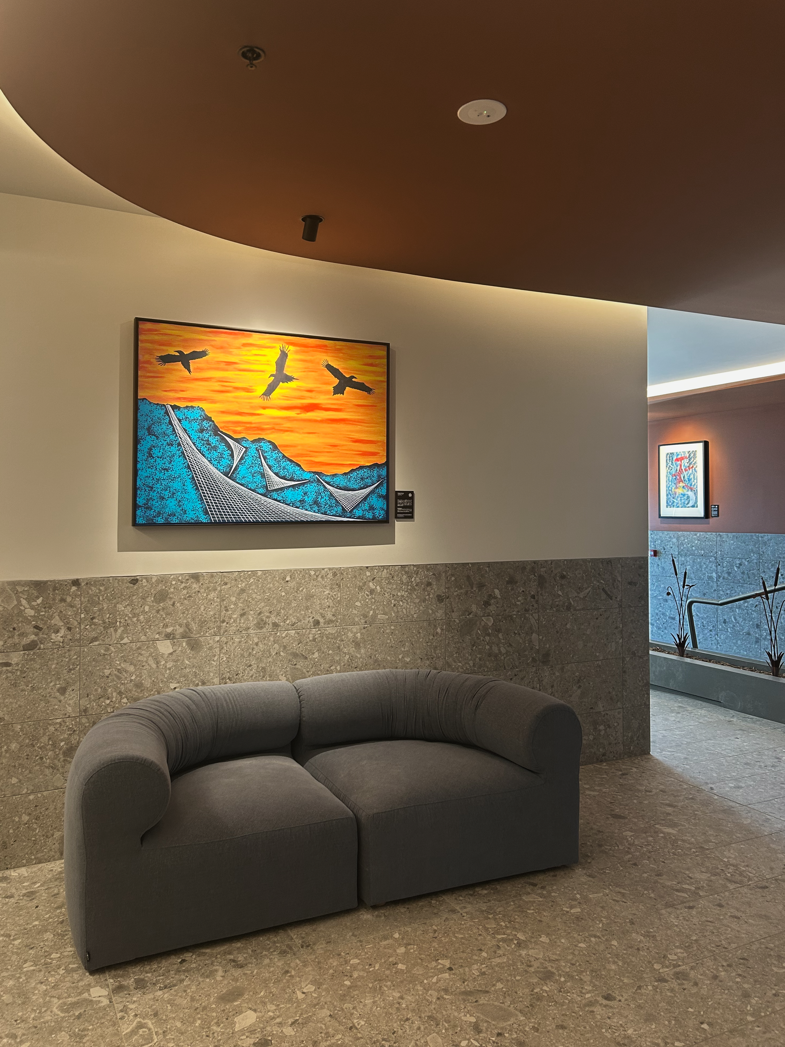 Interior of a building with a gray curved sofa in front of a wall art depicting flying birds and a canyon scene at sunset. There is a framed picture on the adjacent wall and a plant on the floor near a staircase.