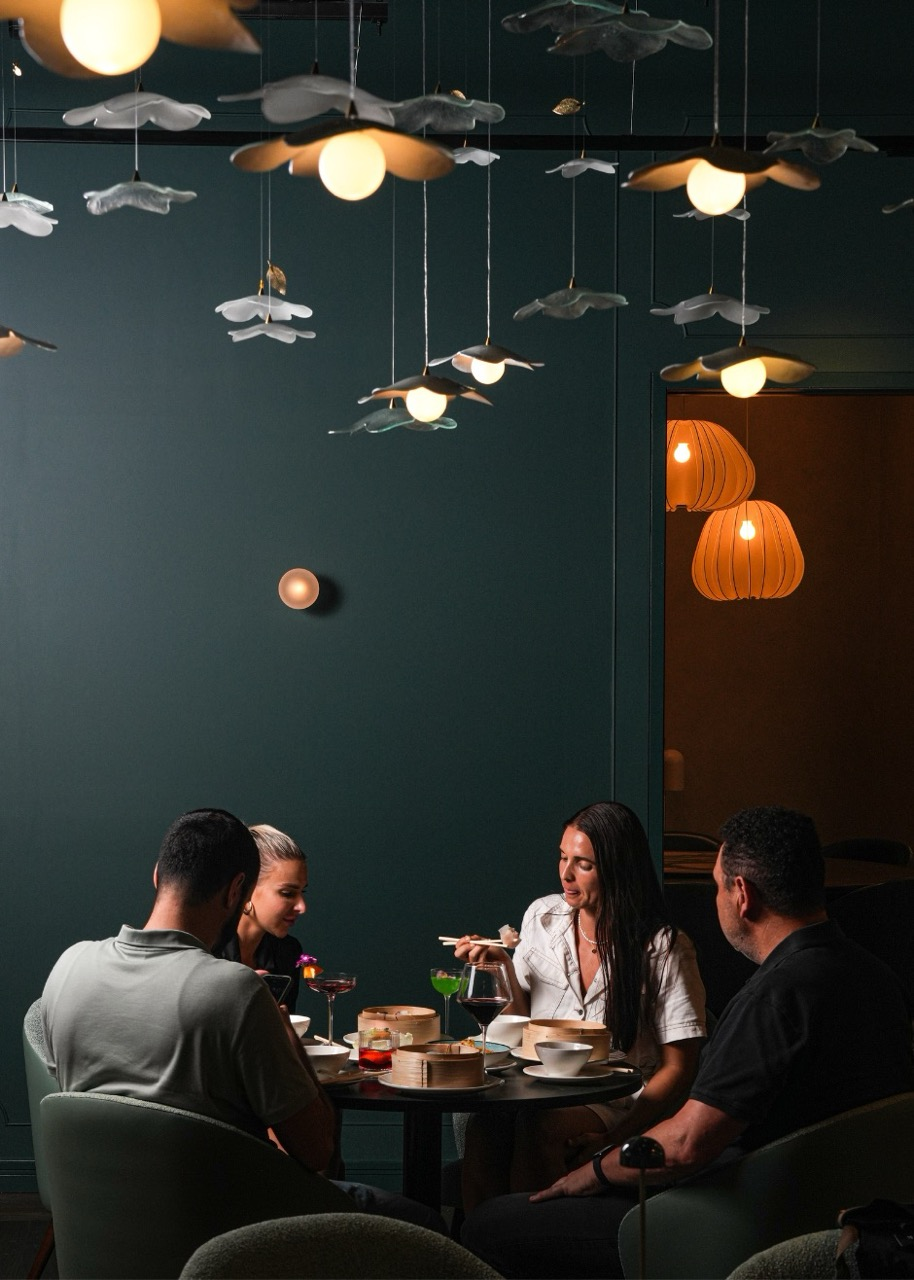 A group of four people seated at a round table in a dimly lit restaurant, with decorative ceiling lights and orange hanging lamps in the background, enjoying a meal with various dishes and drinks.