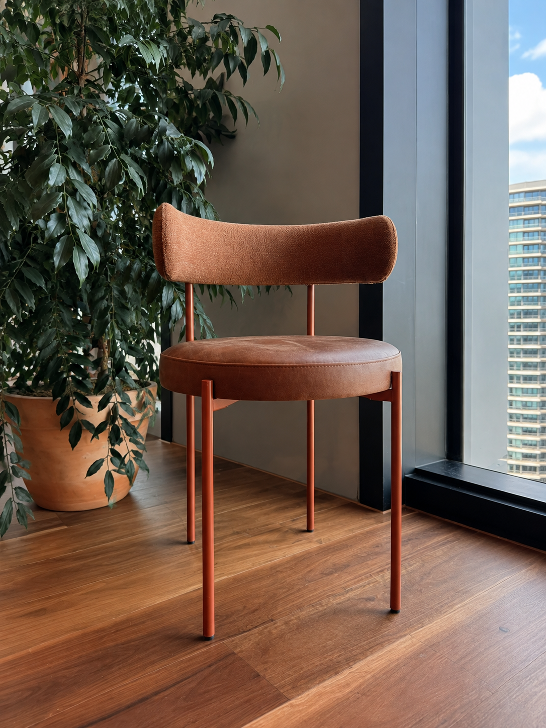 A modern brown chair with a rounded back and thin legs, positioned next to a large window with a view of a city building, with a large green potted plant nearby, on a wooden floor.