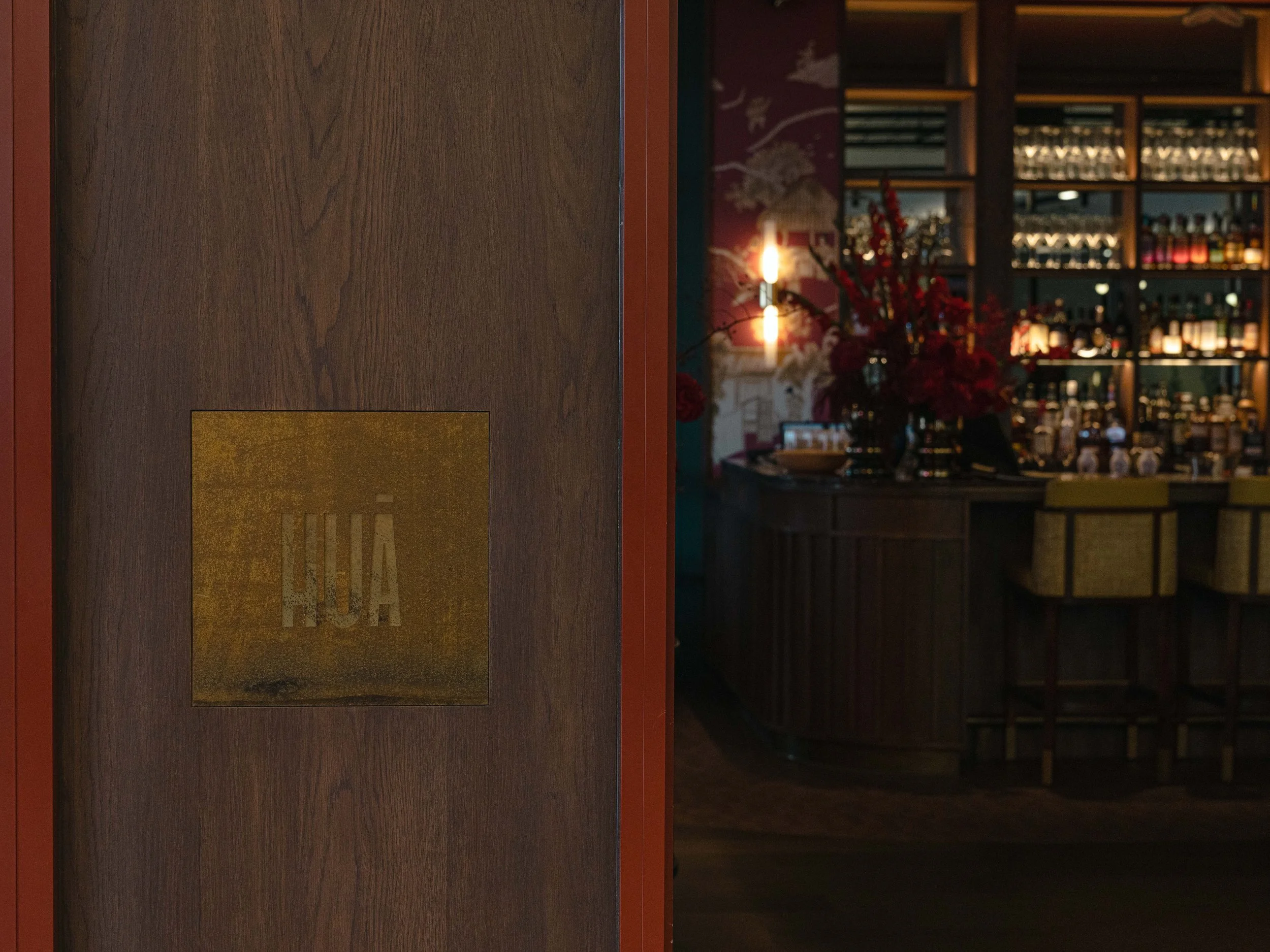 A bar with shelves of liquor bottles, a bouquet of red flowers, and a lit lamp in the background.