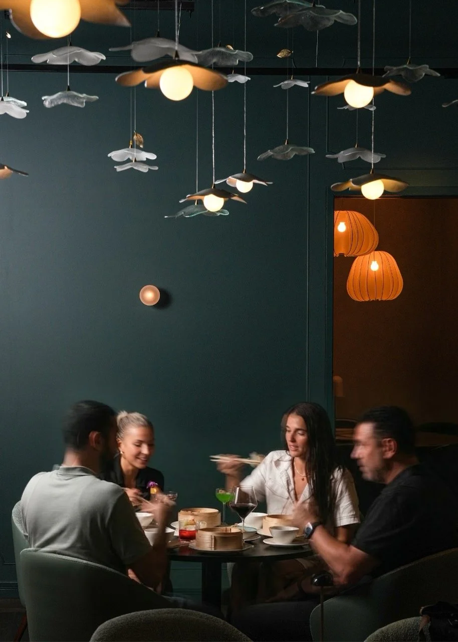 A group of four people seated at a round table in a dimly lit restaurant, with decorative ceiling lights and orange hanging lamps in the background, enjoying a meal with various dishes and drinks.
