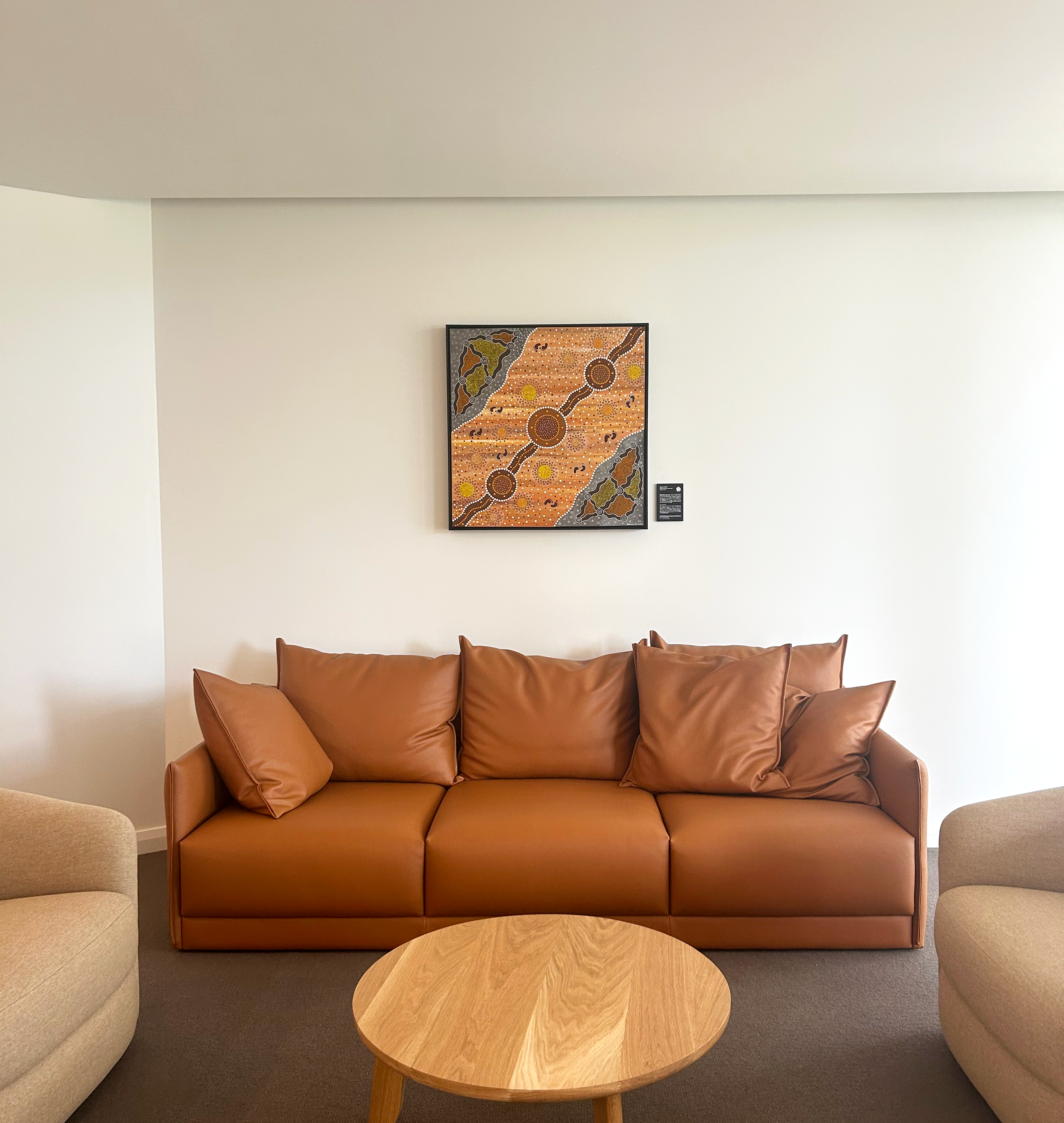 Living room with a brown leather sofa, a wooden coffee table, beige chairs, and an abstract painting on the wall.