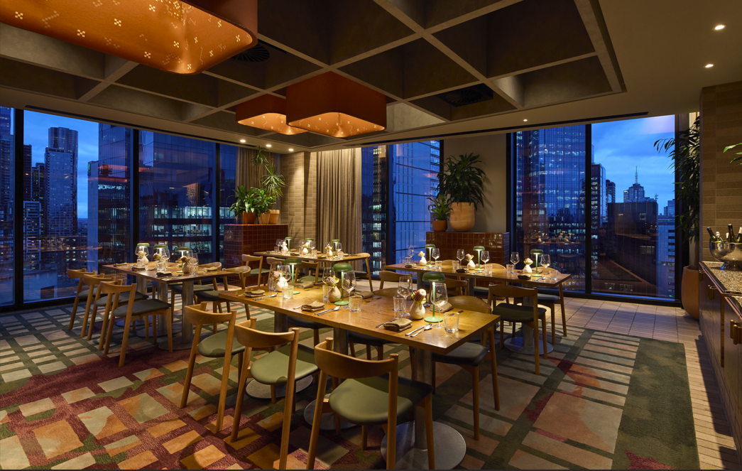 Upscale restaurant dining area with set tables, wine glasses, and flower arrangements, large windows revealing a city skyline at dusk, warm lighting indoors, modern decor with plants and wood accents.