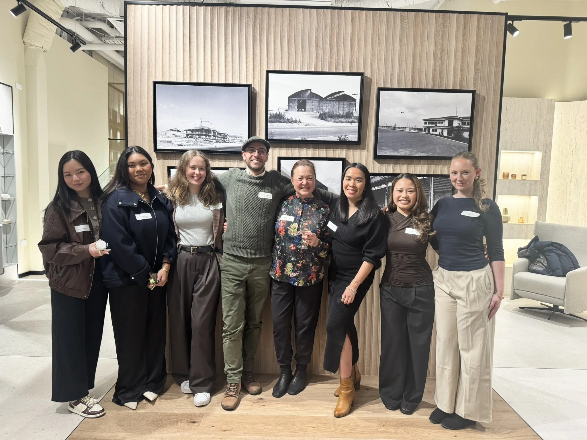 Group of eight people standing together inside a modern gallery or showroom, smiling at the camera. Behind them is a wood-paneled wall with framed black-and-white photos of buildings and construction sites.