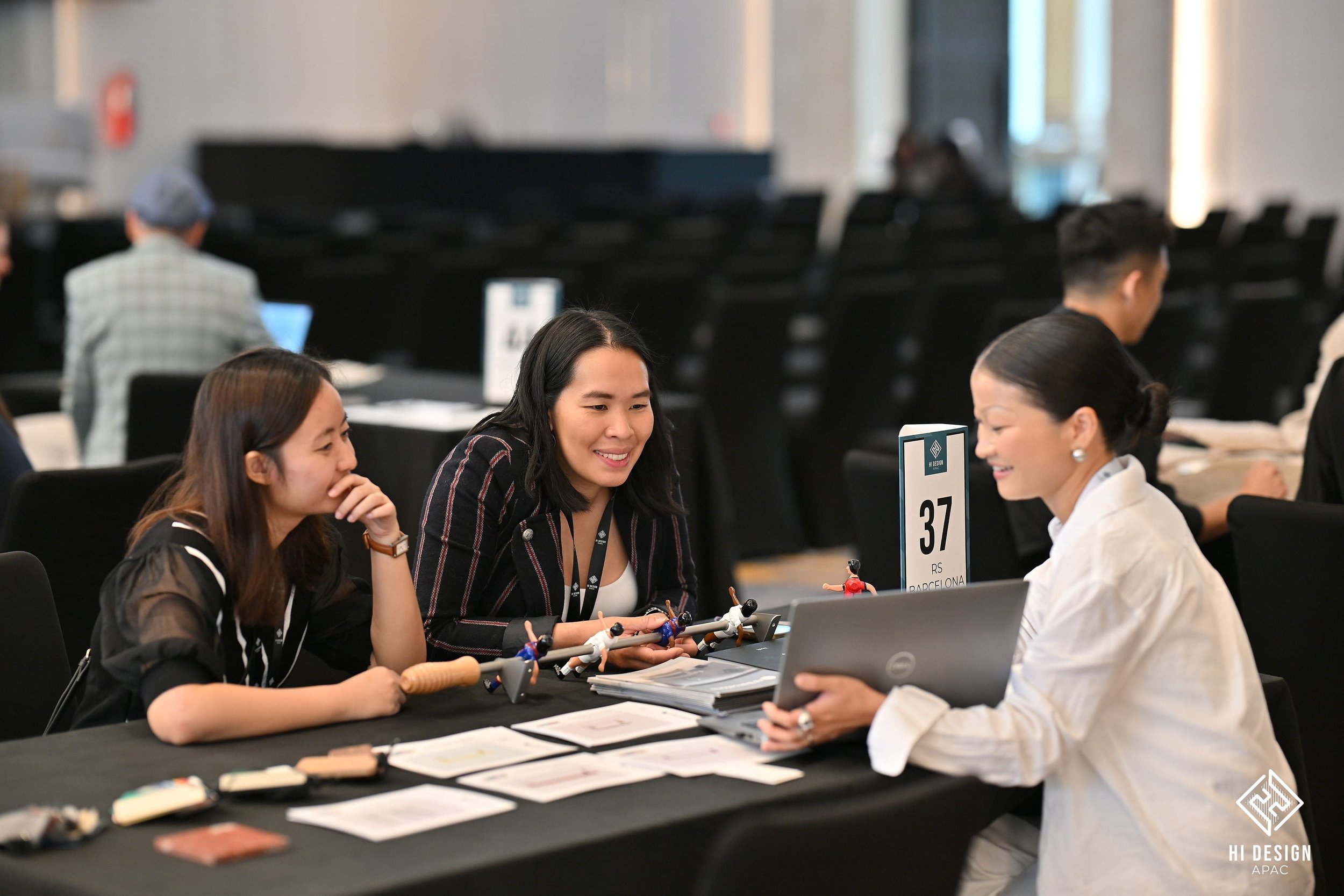 Three women sitting at a table engaged in conversation, with a woman in white speaking and others smiling, at a professional event or conference with numbered signs and materials on the table.