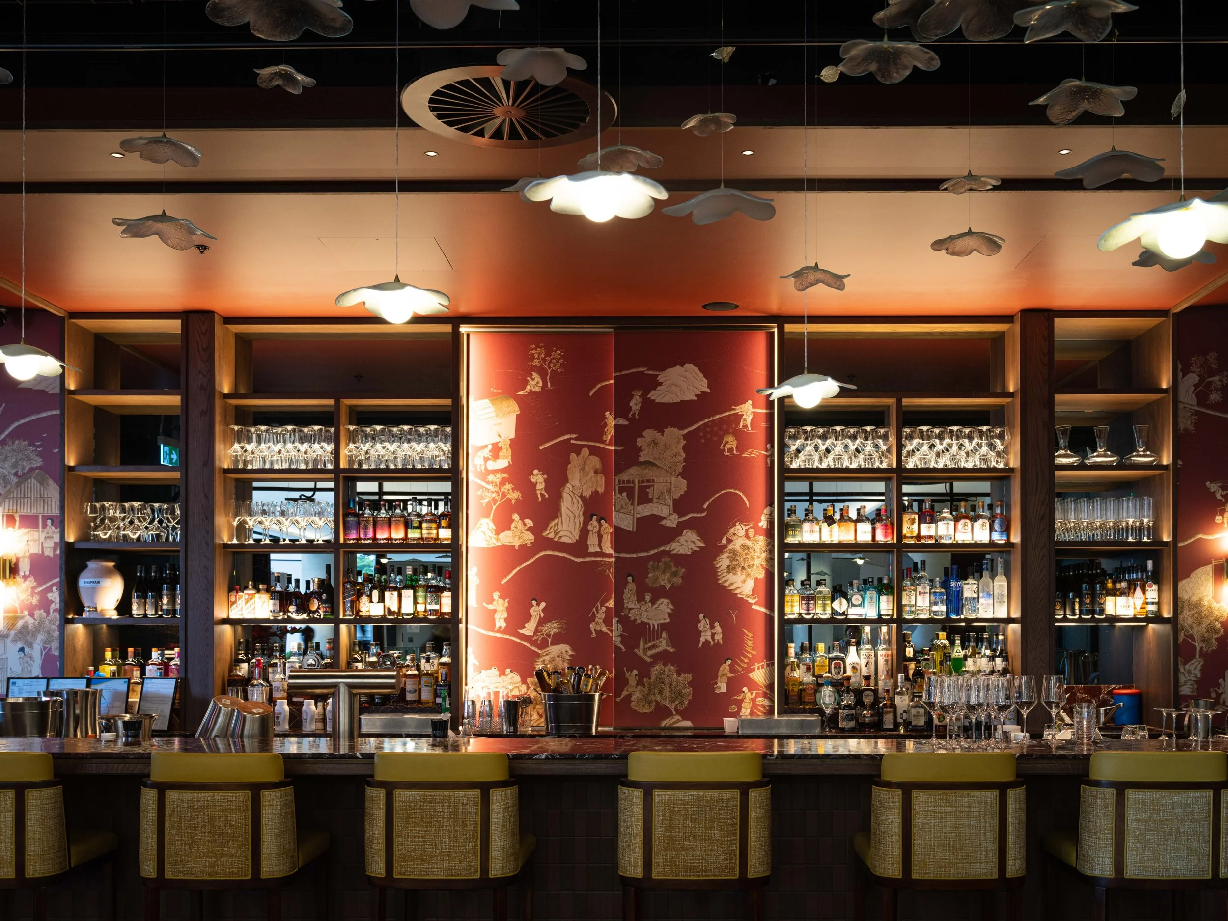Bar with shelves of liquor bottles and glasses, decorated with red wallpaper and hanging white cloud-shaped lamps.