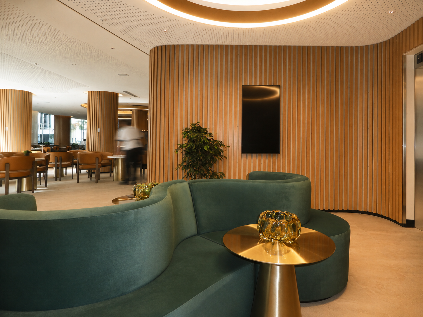 Modern hotel lobby with green curved seating, a small gold table with gold decorative piece, wooden wall paneling, and a potted plant, with part of dining area and a waiter blurred in motion in the background.