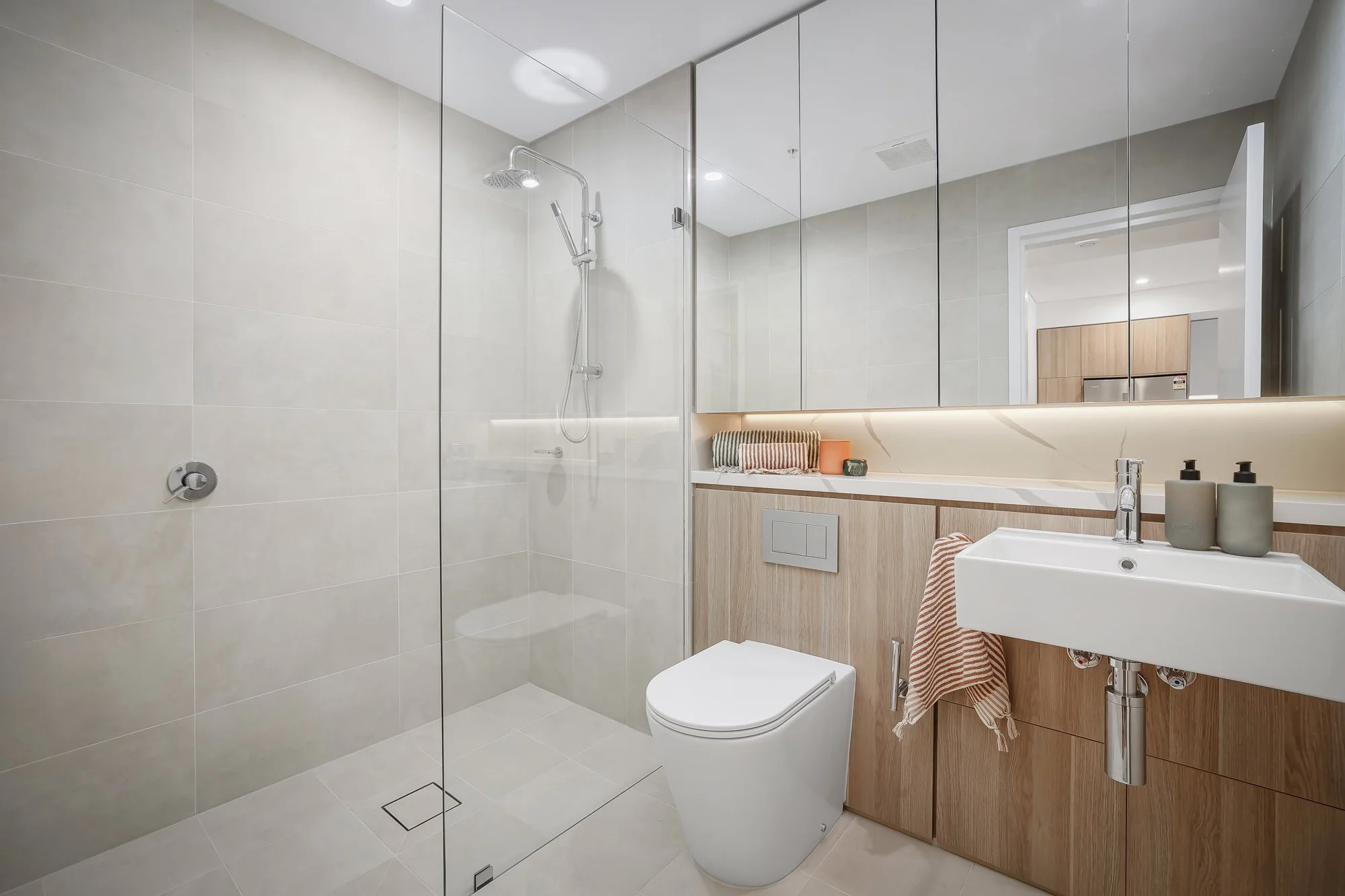 Modern bathroom with a glass-enclosed shower, white toilet, and a wooden vanity with a white sink, large mirrors, and decorative items on the counter.