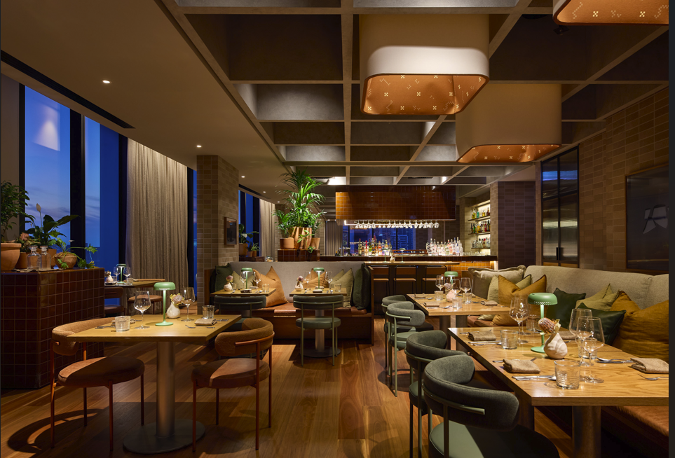 Interior of a modern restaurant bar with wooden tables, upholstered chairs, large windows with curtains, and a well-lit bar area in the background.