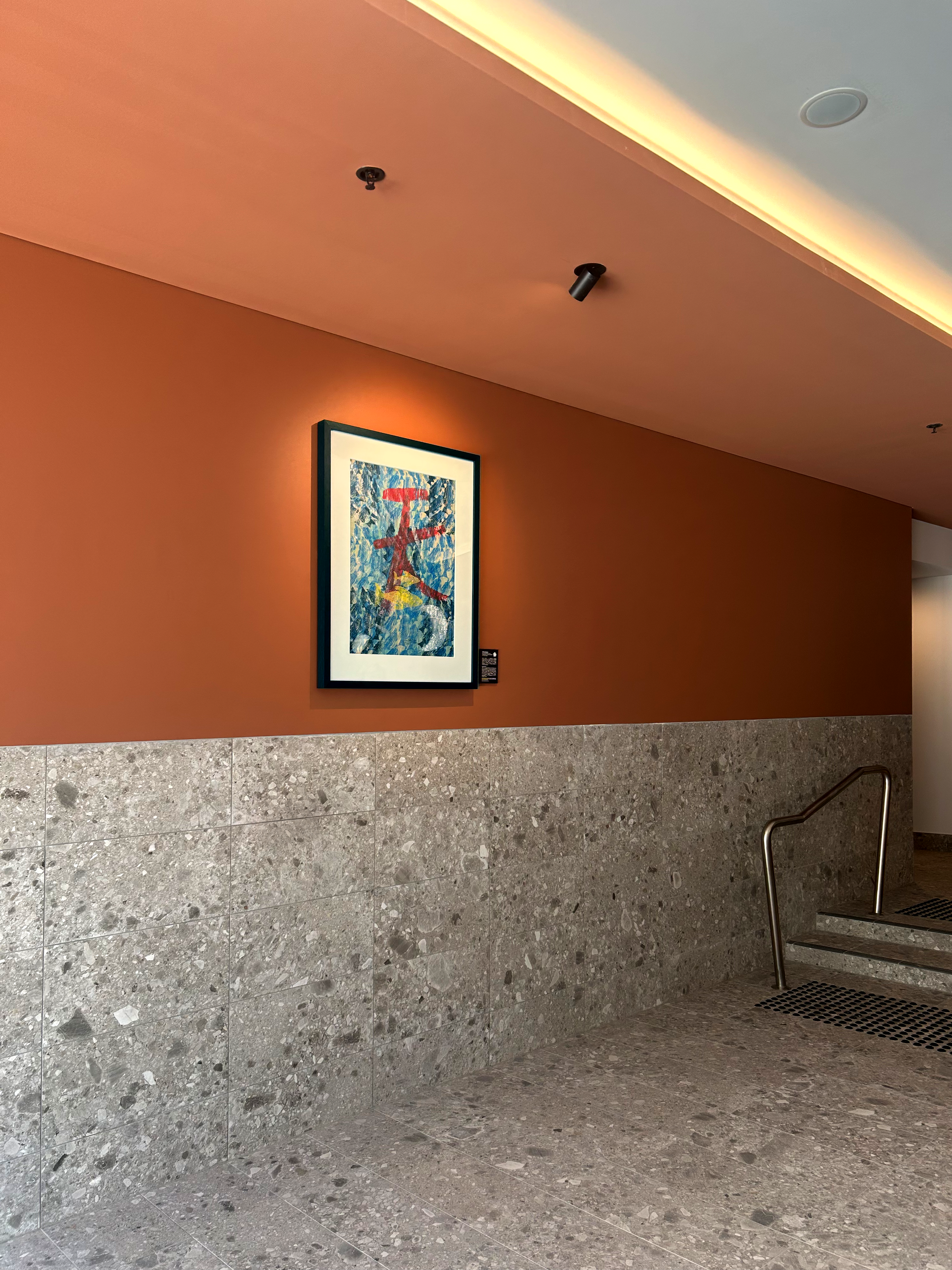 Interior view of a wall with a framed abstract painting, a light-colored ceiling with recessed lighting, a drain grate, and a handrail, with beige and gray tiled flooring and wall. Two ceiling spotlights are visible.