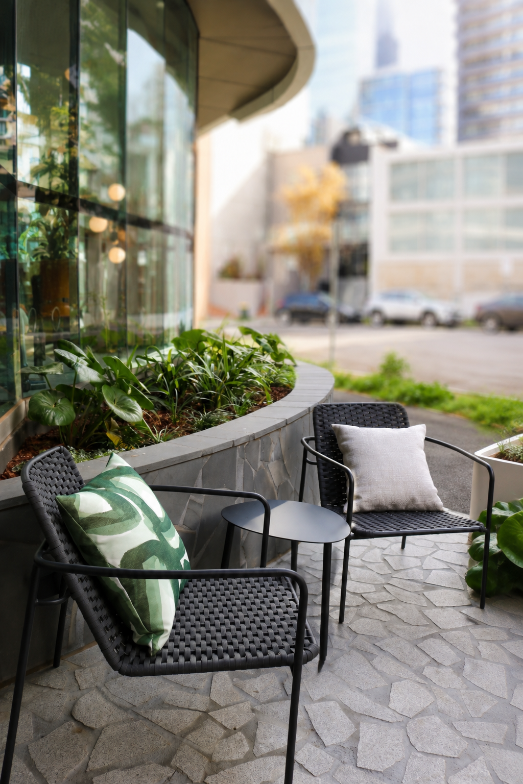 Outdoor patio with two black woven chairs, each with a cushion, and a small round black table between them, near a building with large glass windows and green plants.