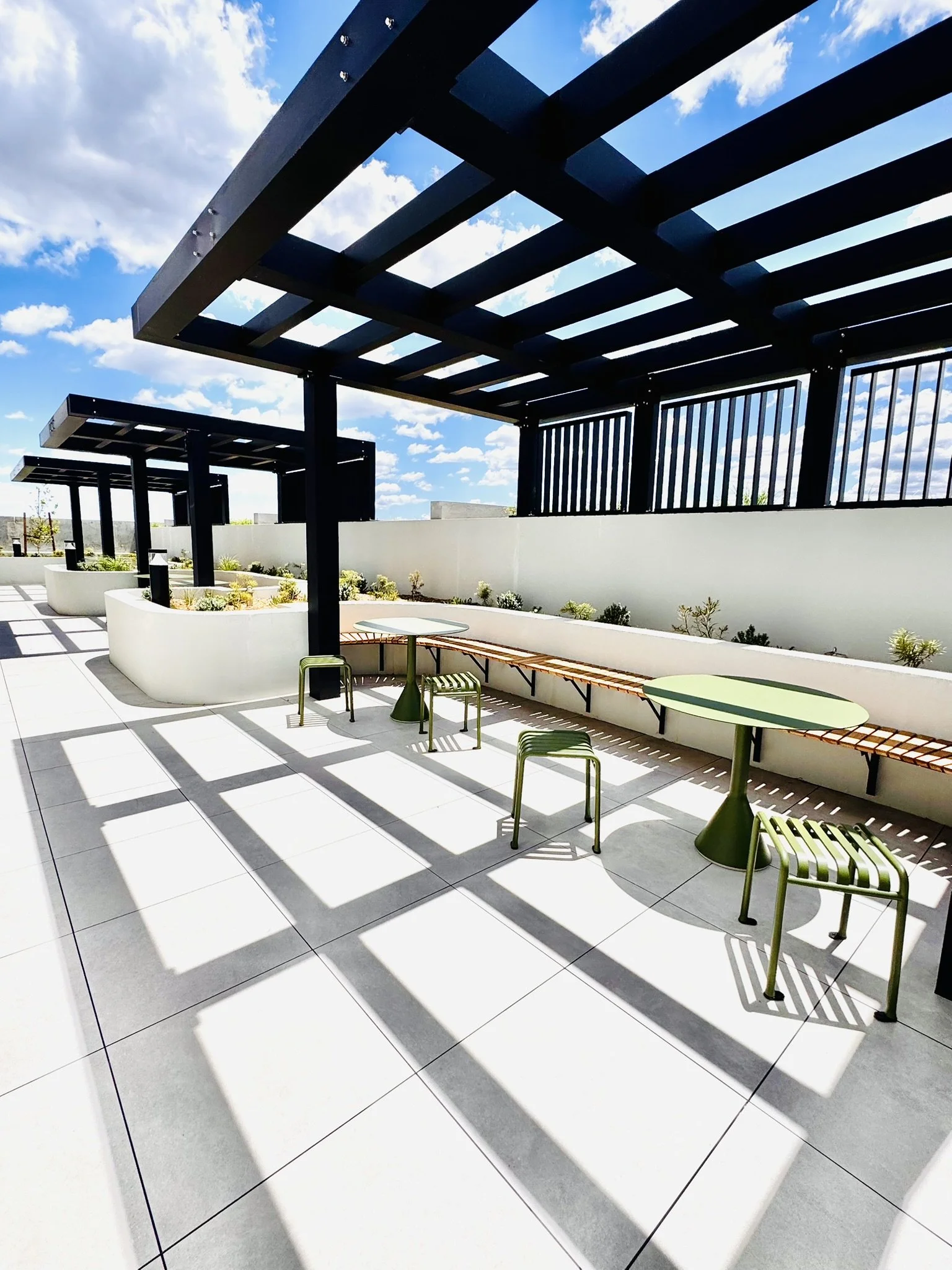 Open outdoor patio area with white tile flooring, green tables, green chairs, wooden bench seating, and black shade structures against a partly cloudy sky.