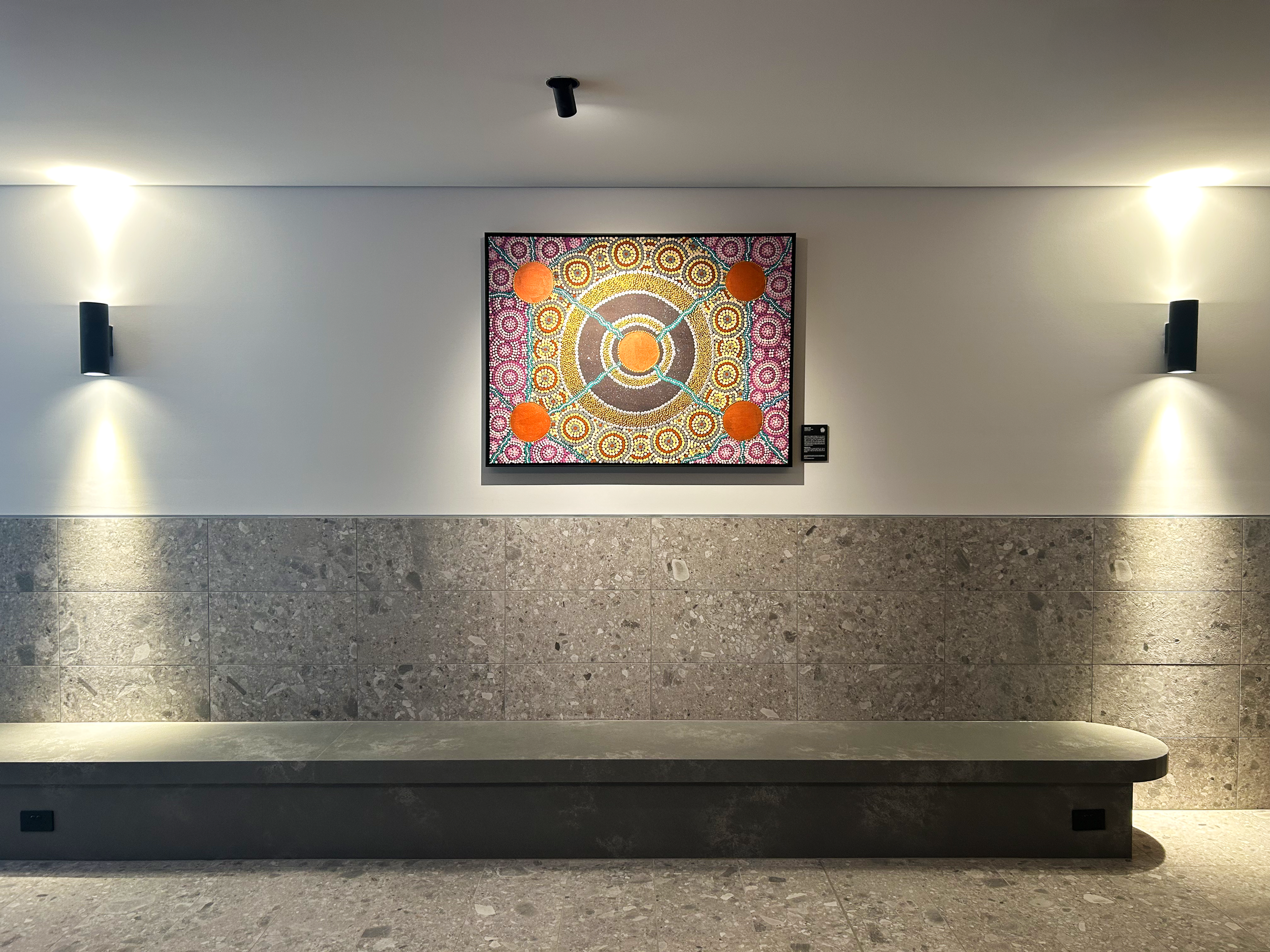 A colorful piece of artwork on a white wall, illuminated by wall-mounted lights on either side, with a textured gray marble floor below.