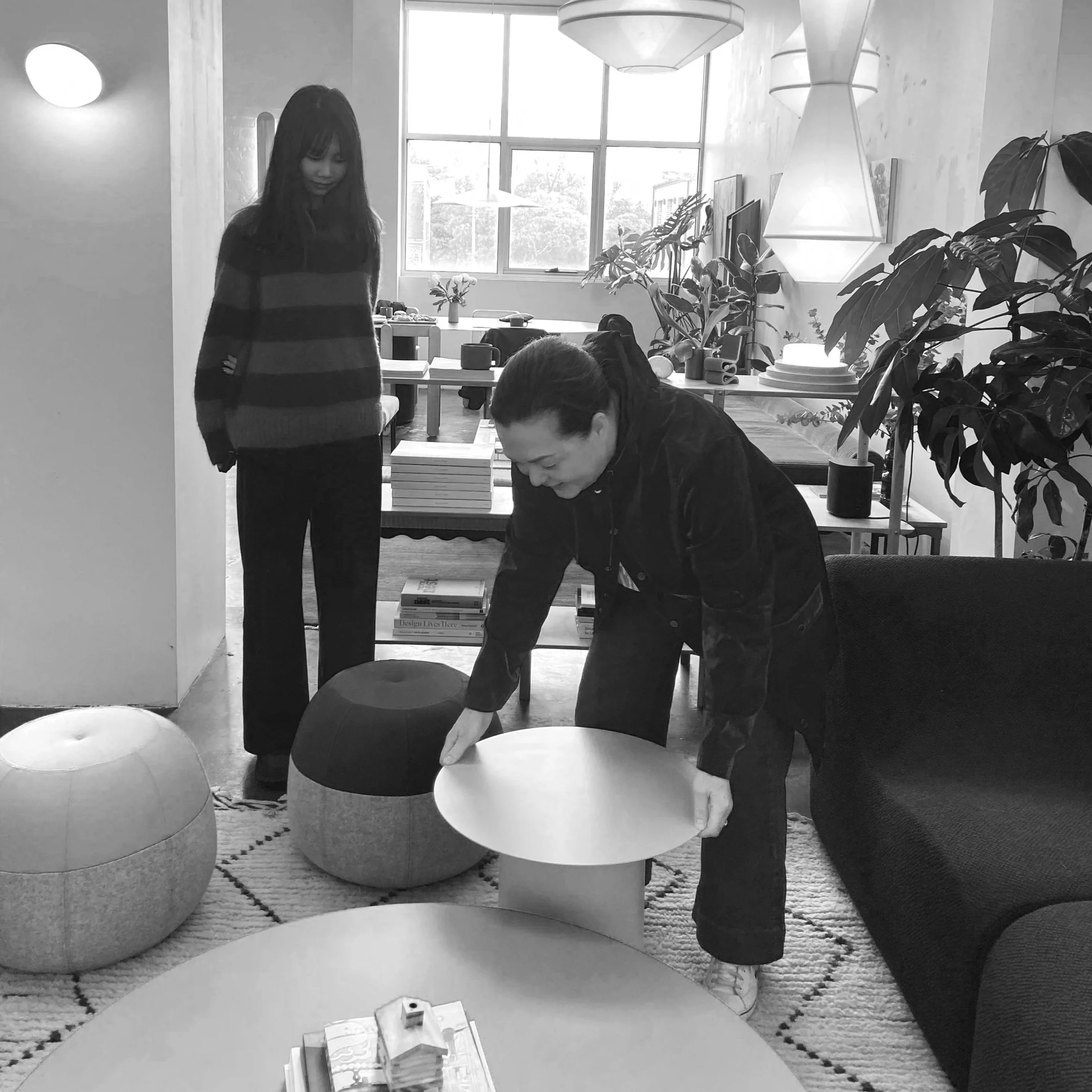 A black and white photo of an indoor space with a woman standing and a man sitting at a table. The room has large windows, plants, and modern light fixtures. The table has books and magazines on it, and there are chairs and a sofa around the room.