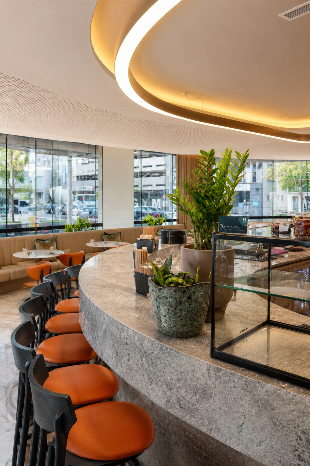 Interior of a modern cafe with a curved marble counter, several orange and black chairs, potted plants, and large windows showing trees and parked cars outside.