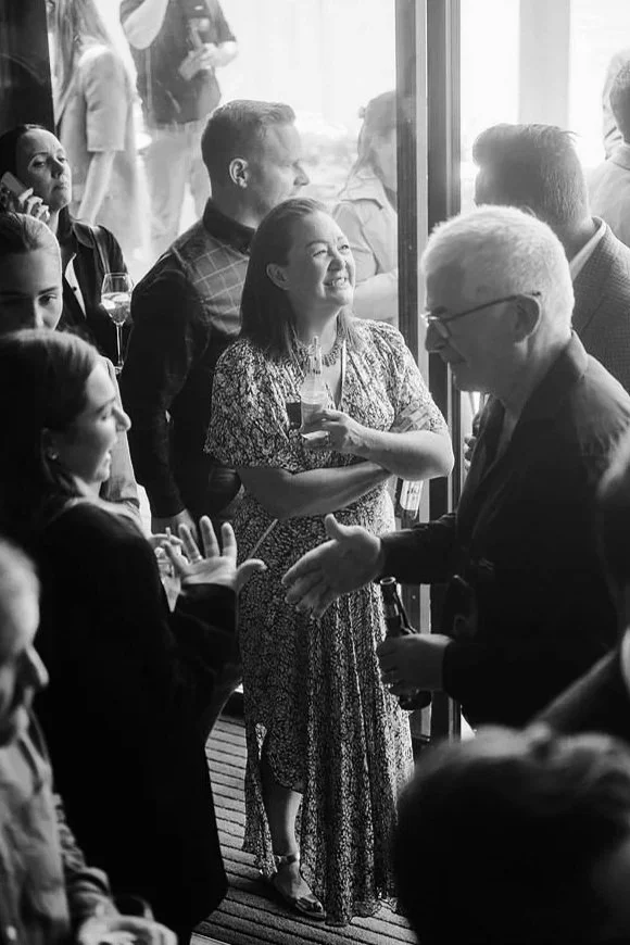 People socializing at a gathering, including a woman smiling holding a drink, and a man with glasses talking to another person, with others in the background.