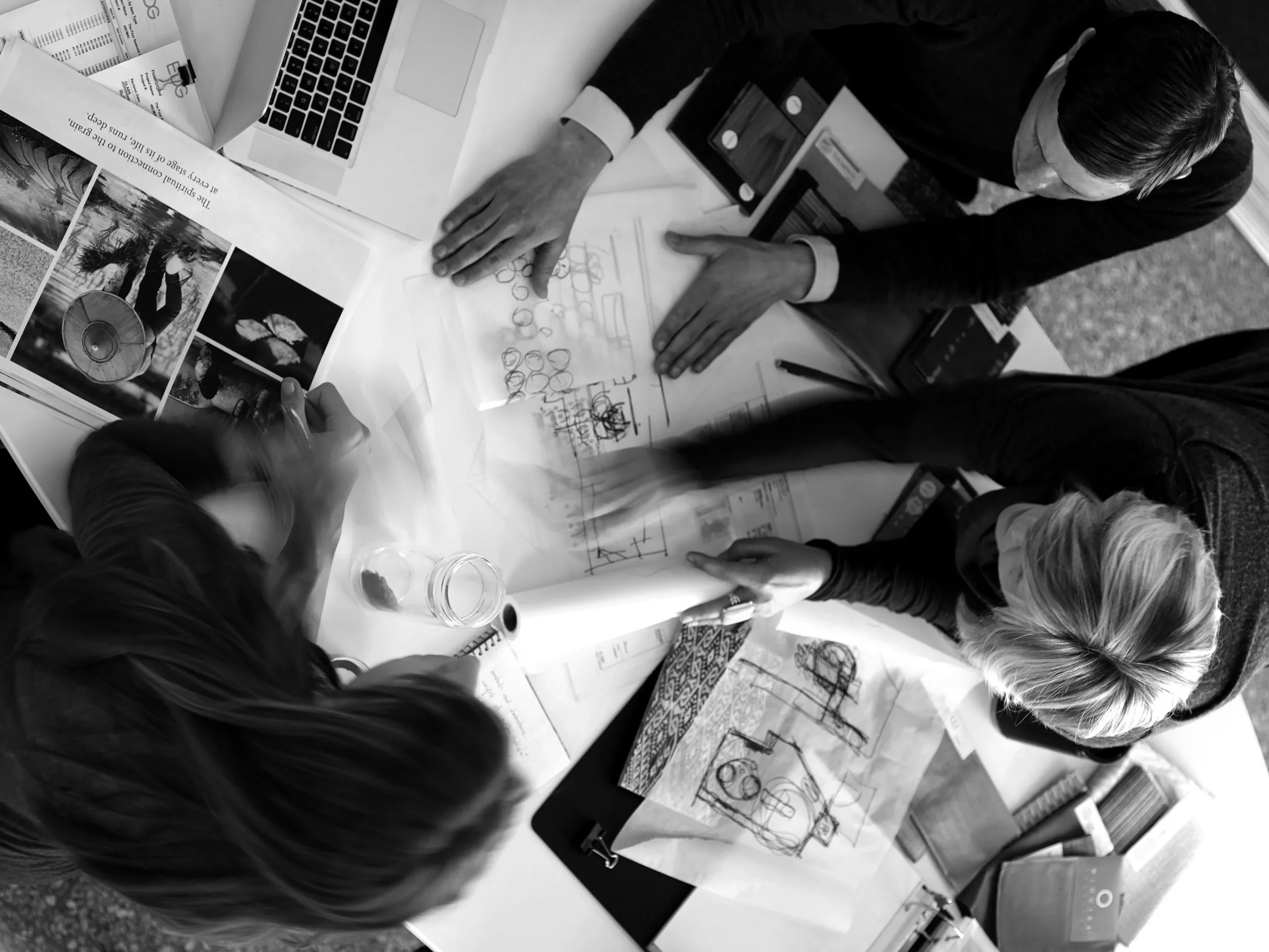 Three people are gathered around a table working on architectural or design plans, with various sketches, photos, and documents spread out on the table, and a laptop is also visible.