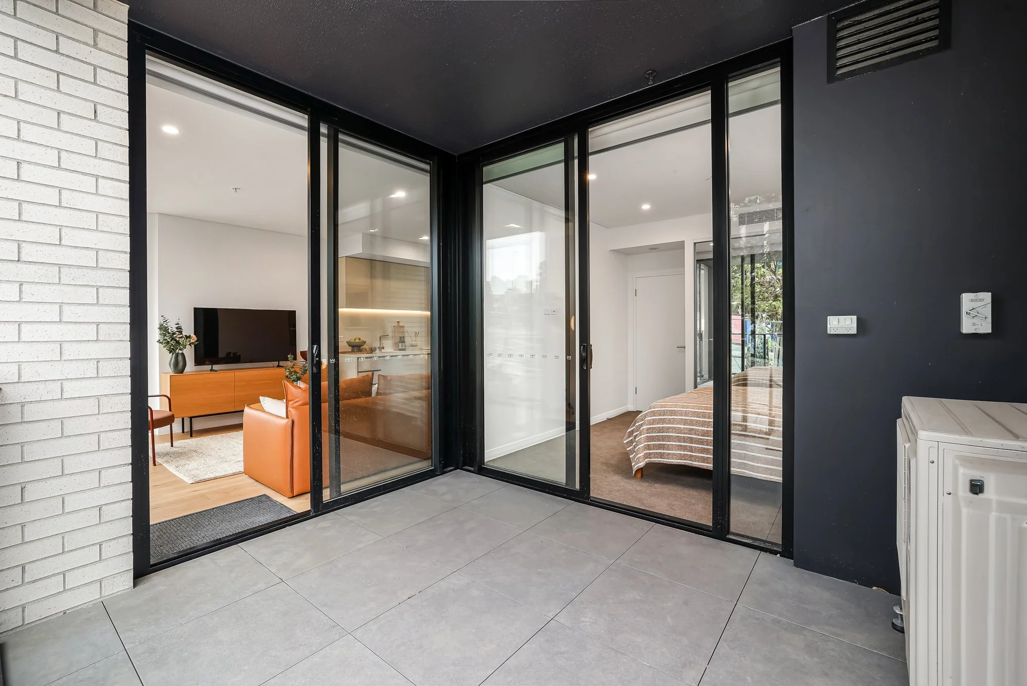 View from an outdoor balcony into a living room and bedroom separated by glass sliding doors. The living room has a TV on a wooden cabinet and an orange armchair; the bedroom has a bed with a striped brown and white blanket. The balcony has grey tile