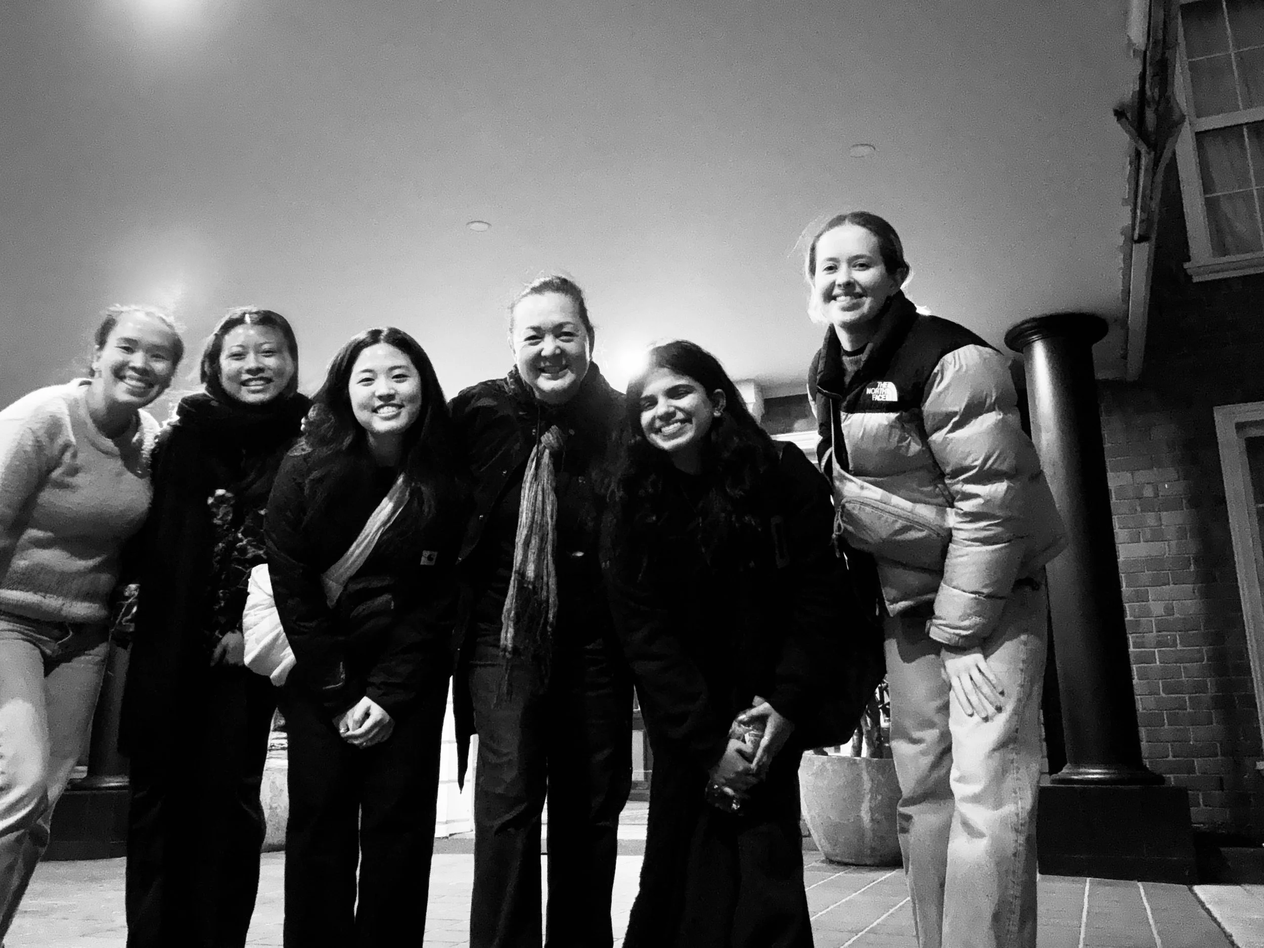 Group of six smiling people standing together indoors, with a brick wall and furniture in the background.