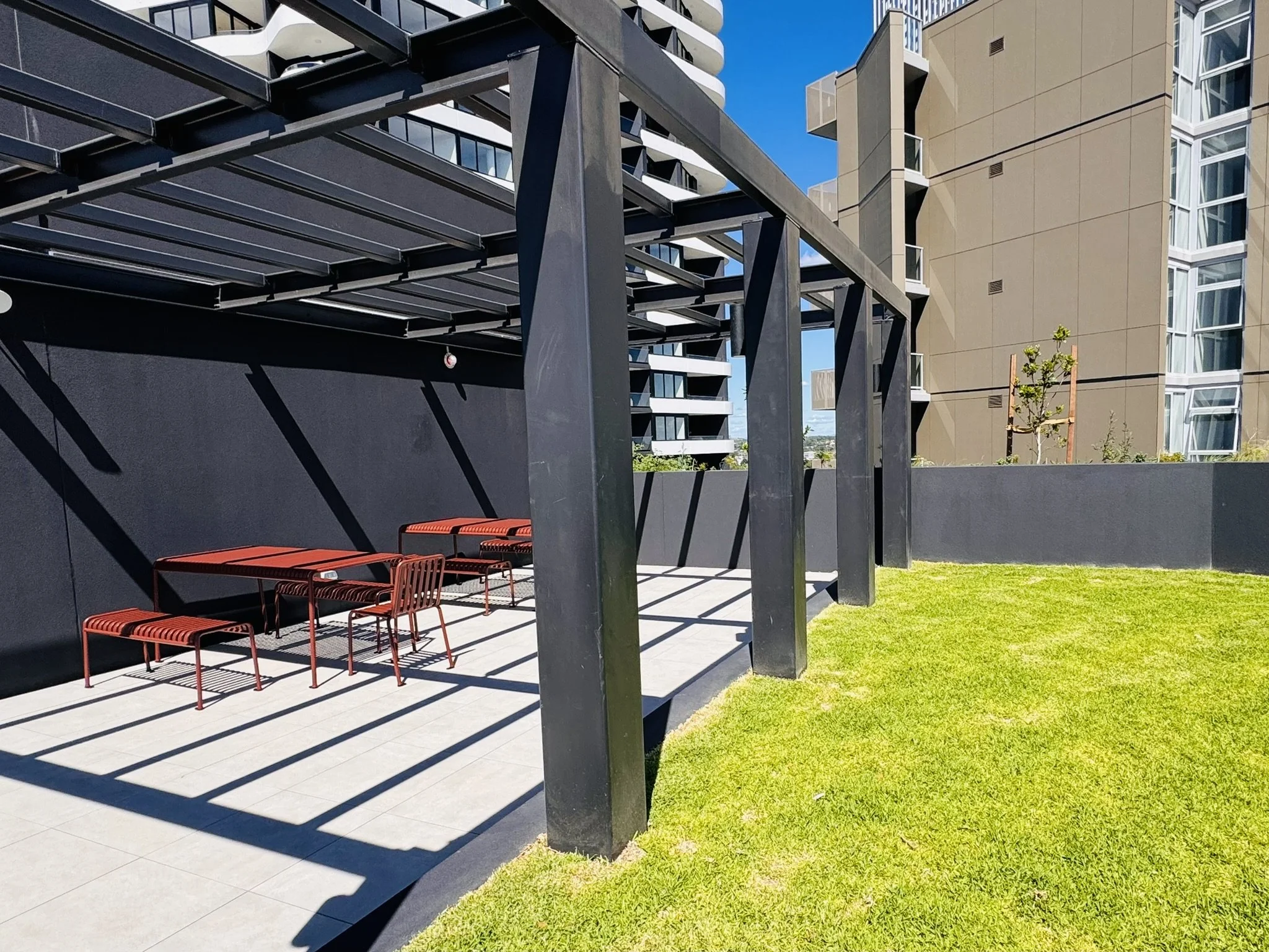 An outdoor patio area with a black metal pergola, red chairs with striped cushions, and a black wall. There is a grassy lawn in the foreground and modern apartment buildings in the background under a clear blue sky.