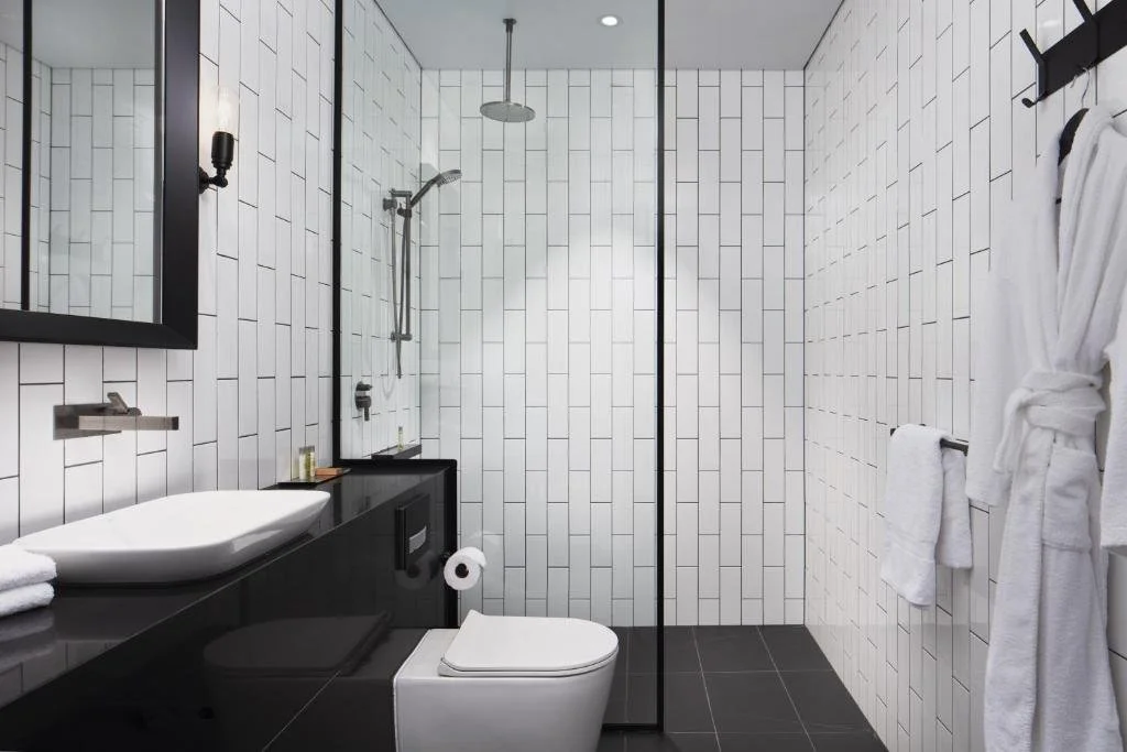 Modern bathroom with white tiled walls, black vanity with vessel sink, toilet, walk-in shower with glass partition, and white towels hanging on wall