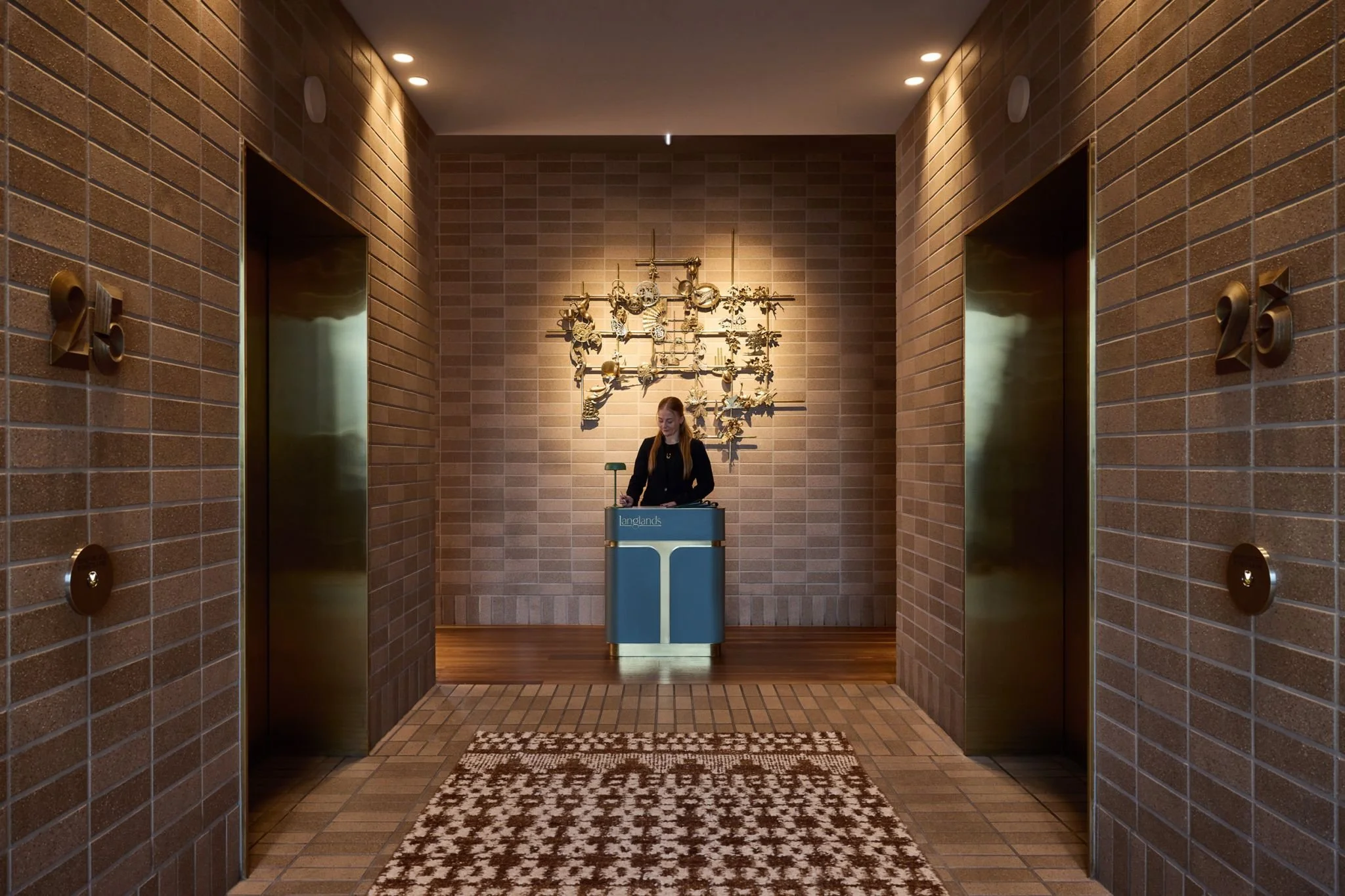 A woman in black standing behind a reception desk in a hallway with brown brick walls and two elevators.