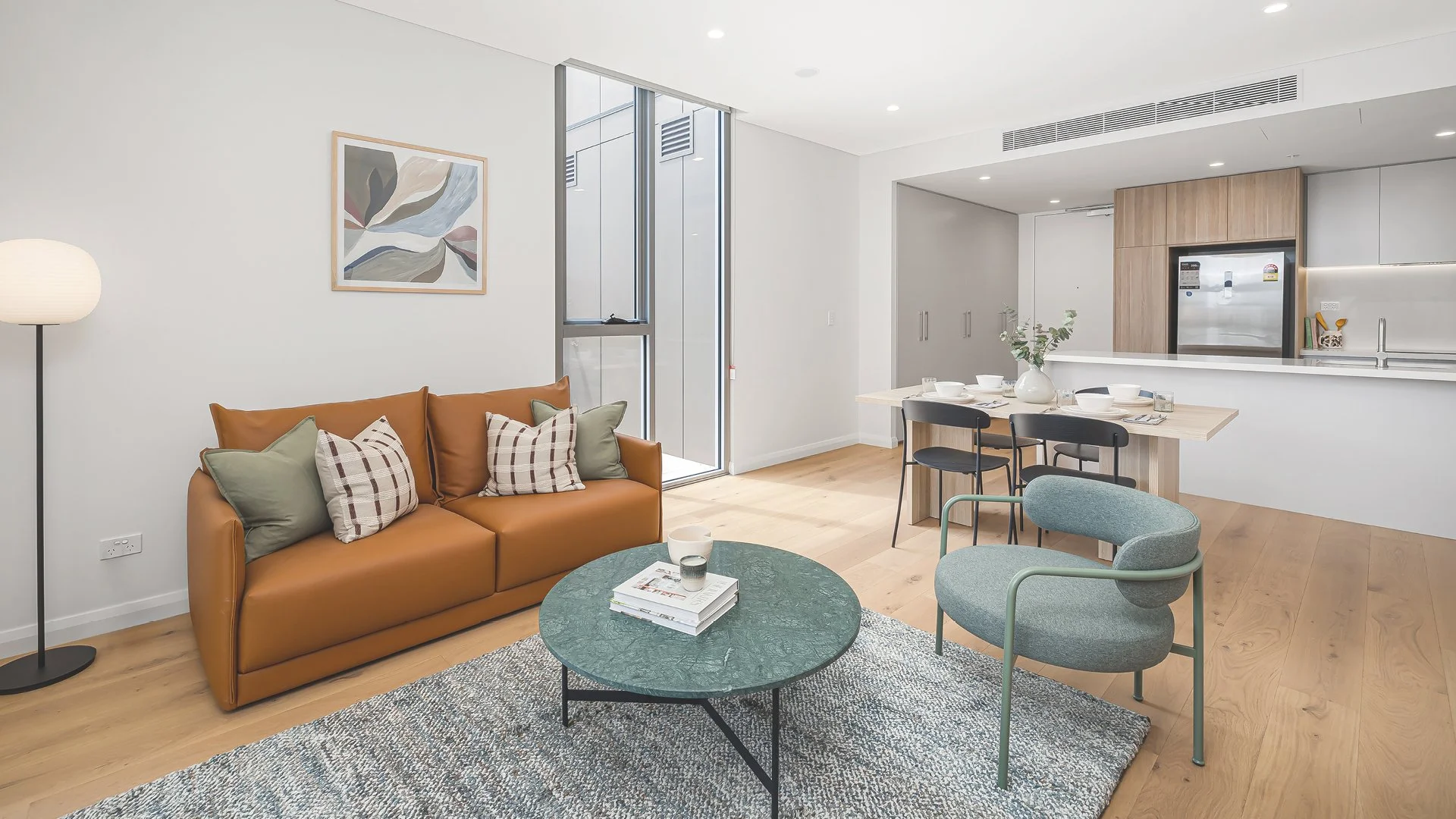 Modern living room with a tan leather sofa, decorative cushions, a green marble coffee table, a grey armchair, a white dining table set, and a kitchen area in the background with wood and stainless steel finishes.