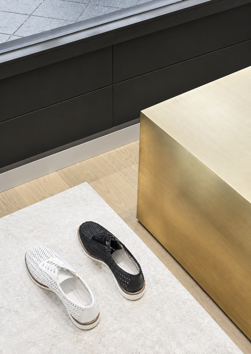 A pair of black and white woven slip-on shoes on a white marble surface next to a gold-colored box and black cabinetry with tiled floor in the background.