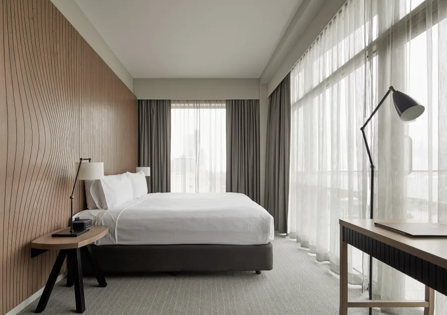 A modern hotel room with a bed covered in white linens, a bedside table with a lamp, and a large window with sheer curtains and blackout drapes.
