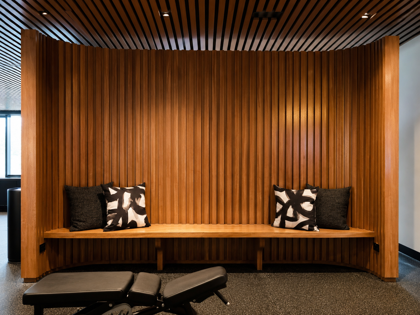 Modern wooden seating area with black and white patterned pillows, against a curved vertical wood-paneled wall in a contemporary interior space.
