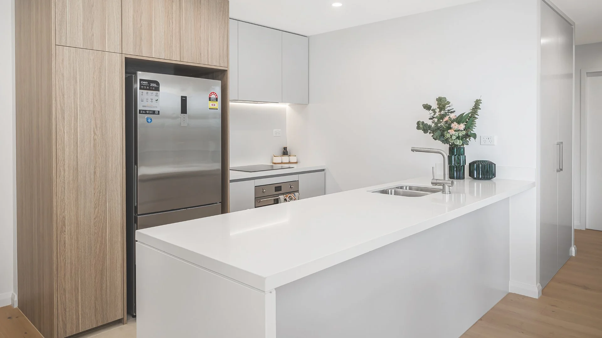 Modern kitchen with a white countertop, a stainless steel refrigerator, built-in oven, and minimalist decor including a vase of flowers.