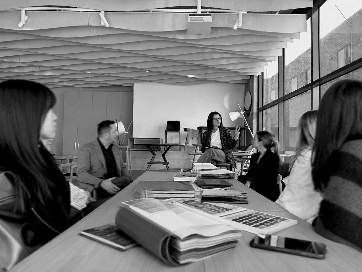 A business meeting taking place in a modern conference room with large windows and a rectangular table surrounded by attendees. One woman is presenting, sitting on a higher platform, gesturing with her hand. The conference table has documents, magazines, and electronic devices on it.