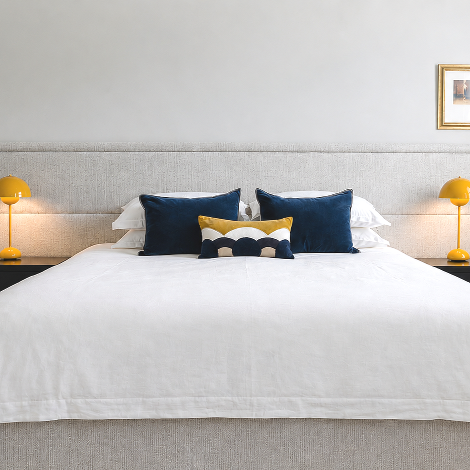 A neatly made bed with white bedding, two dark blue pillows, and a decorative pillow with yellow, navy, and white colors, flanked by yellow bedside lamps on either side.
