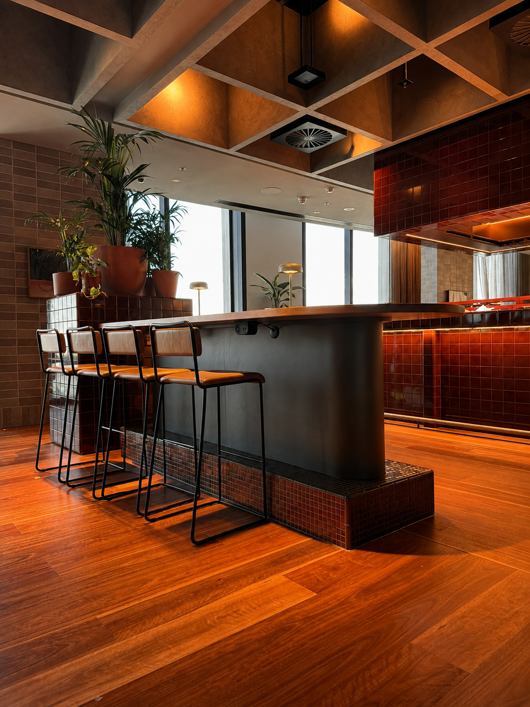 Modern bar area with wooden flooring, a curved bar counter with dark red tiles, and high chairs with wooden seats. There are potted plants on the bar and large windows letting in natural light.