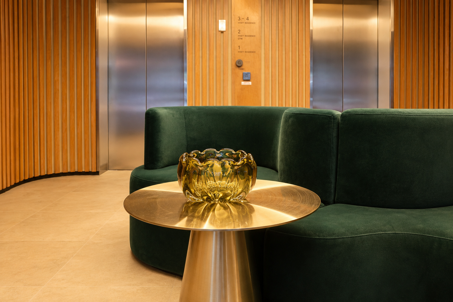 A lobby seating area with a green velvet sofa, a round gold table, and a decorative gold glass bowl. The background features wooden wall panels and two elevator doors.