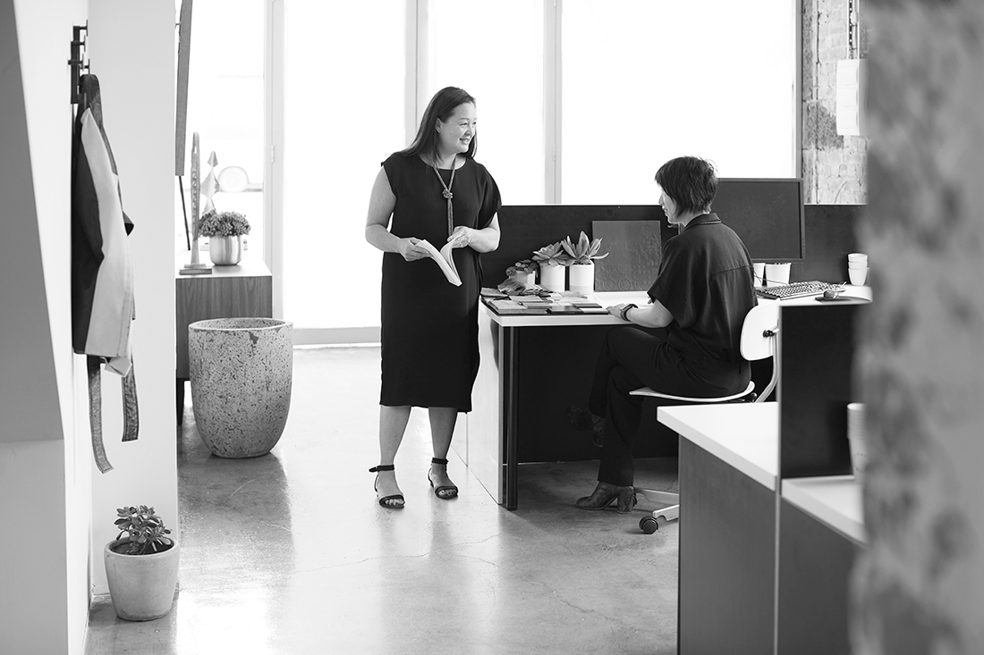 Two women in an office, one standing with a book and the other seated at a desk, engaged in conversation.