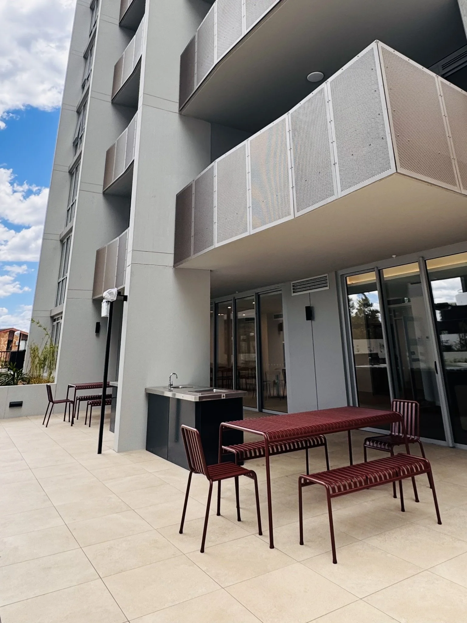 Modern apartment balcony with red patio furniture and outdoor sink, overlooking a city skyline with blue sky and clouds.