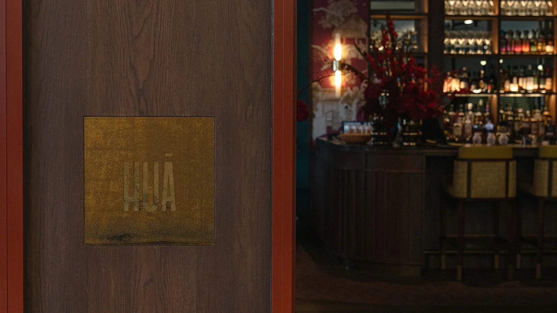 Close-up of a wooden door with a small gold-colored sign that has the word 'NO' on it. In the background, part of a bar with bottles, glasses, and a large floral arrangement is visible.