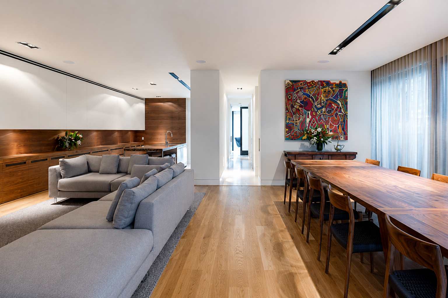 Open-concept living and dining area with wooden flooring, a large wooden dining table with eight chairs, a grey sofa, artwork on the wall, and large windows with sheer curtains.