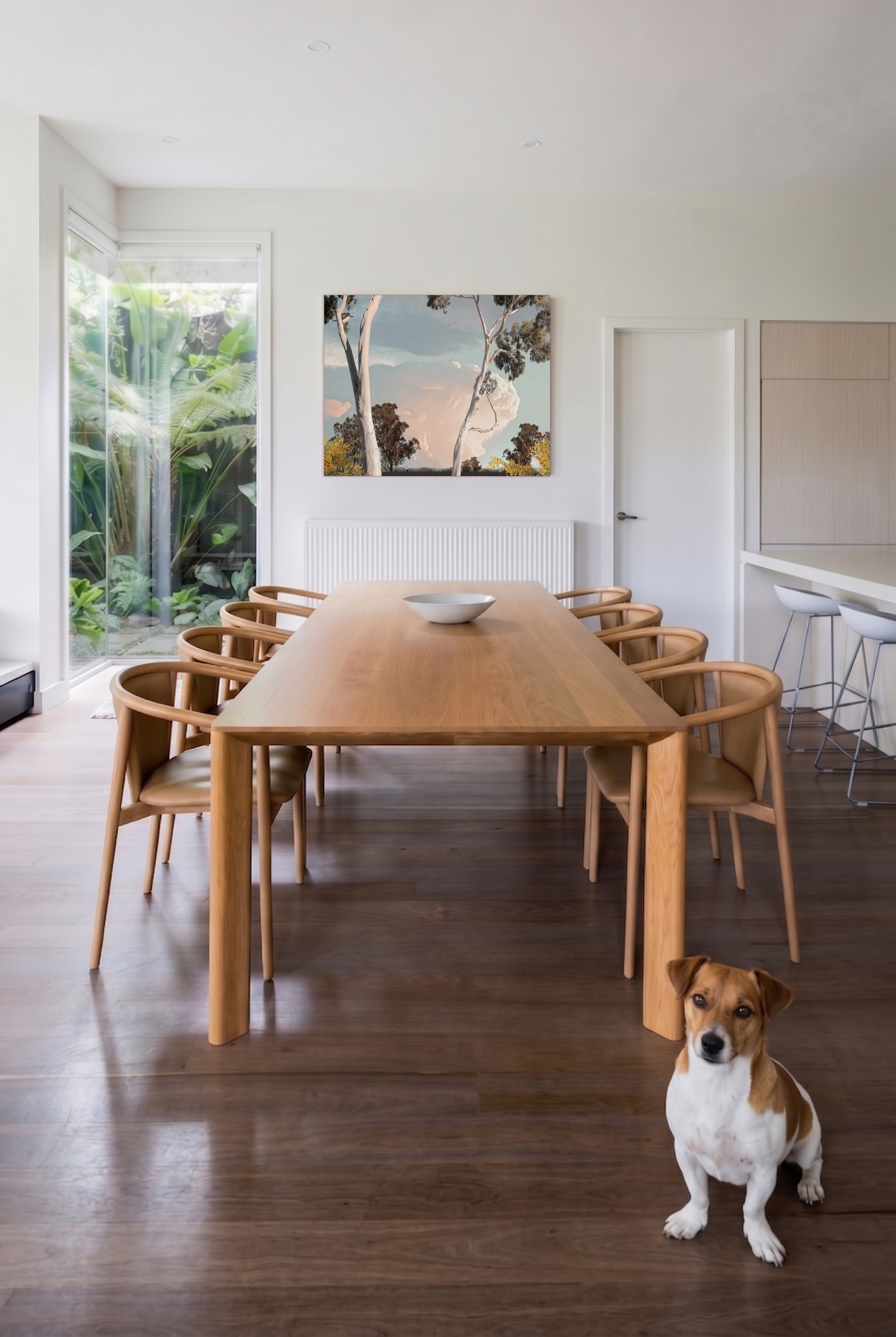 NUMI COLLECTIVE A dining room with a wooden table, chairs, a colorful abstract painting on the wall, a sliding glass door with greenery outside, and a small dog sitting on the floor.