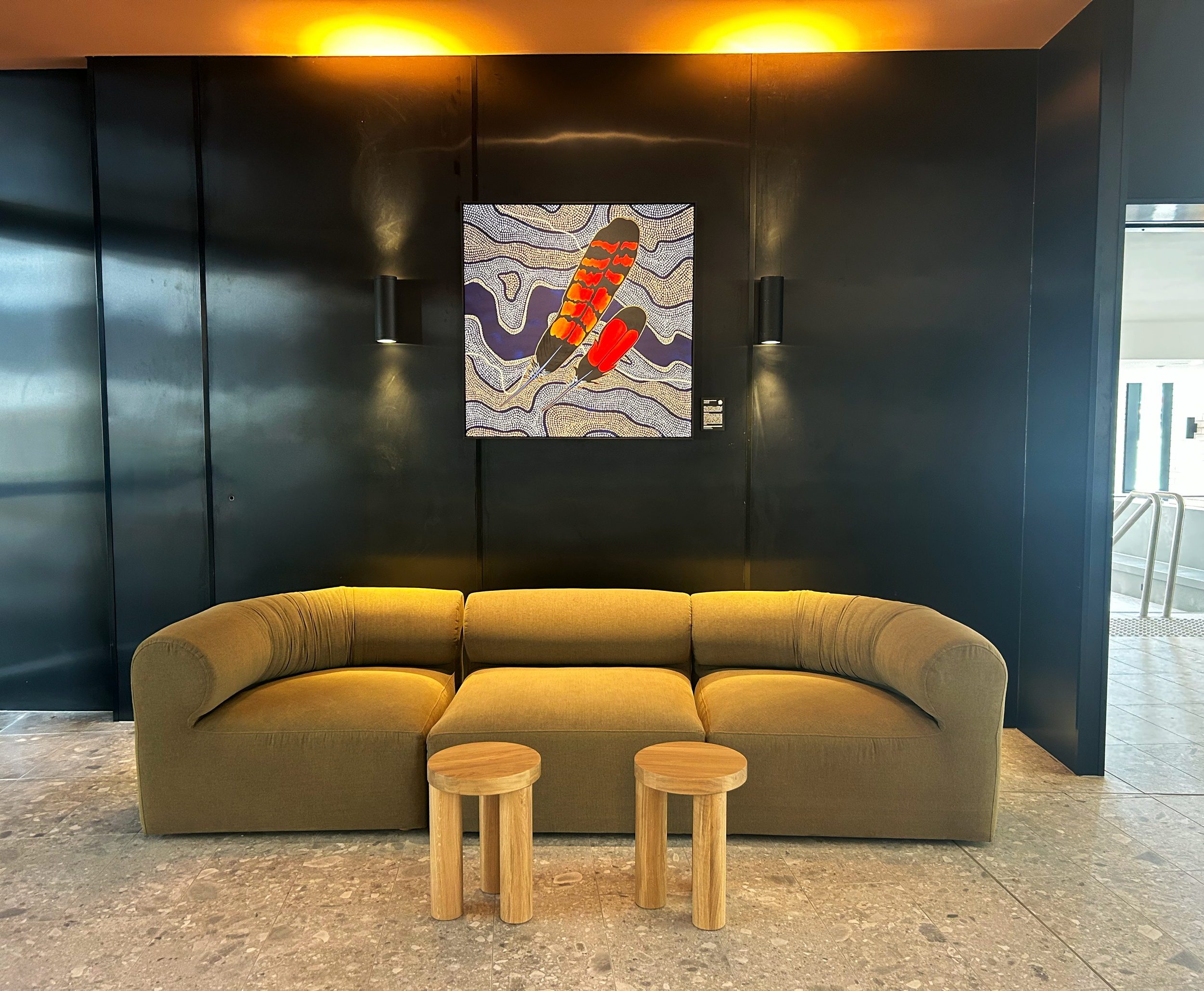 A mustard-colored sofa with two small wooden stools in front of it, situated in a modern interior with dark walls and artistic artworks illuminated with warm overhead lighting.