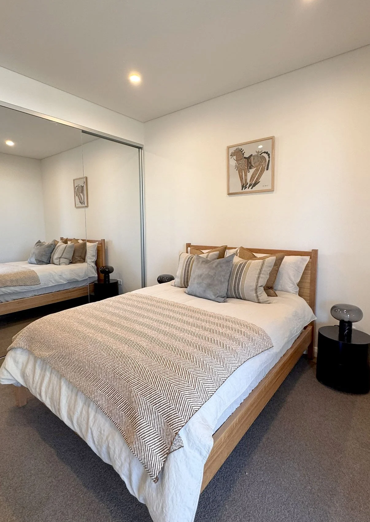 A bedroom with a wooden bed frame and multiple pillows, a patterned blanket, bedside tables with modern lamps, a framed animal artwork on the wall, and a mirrored closet door.