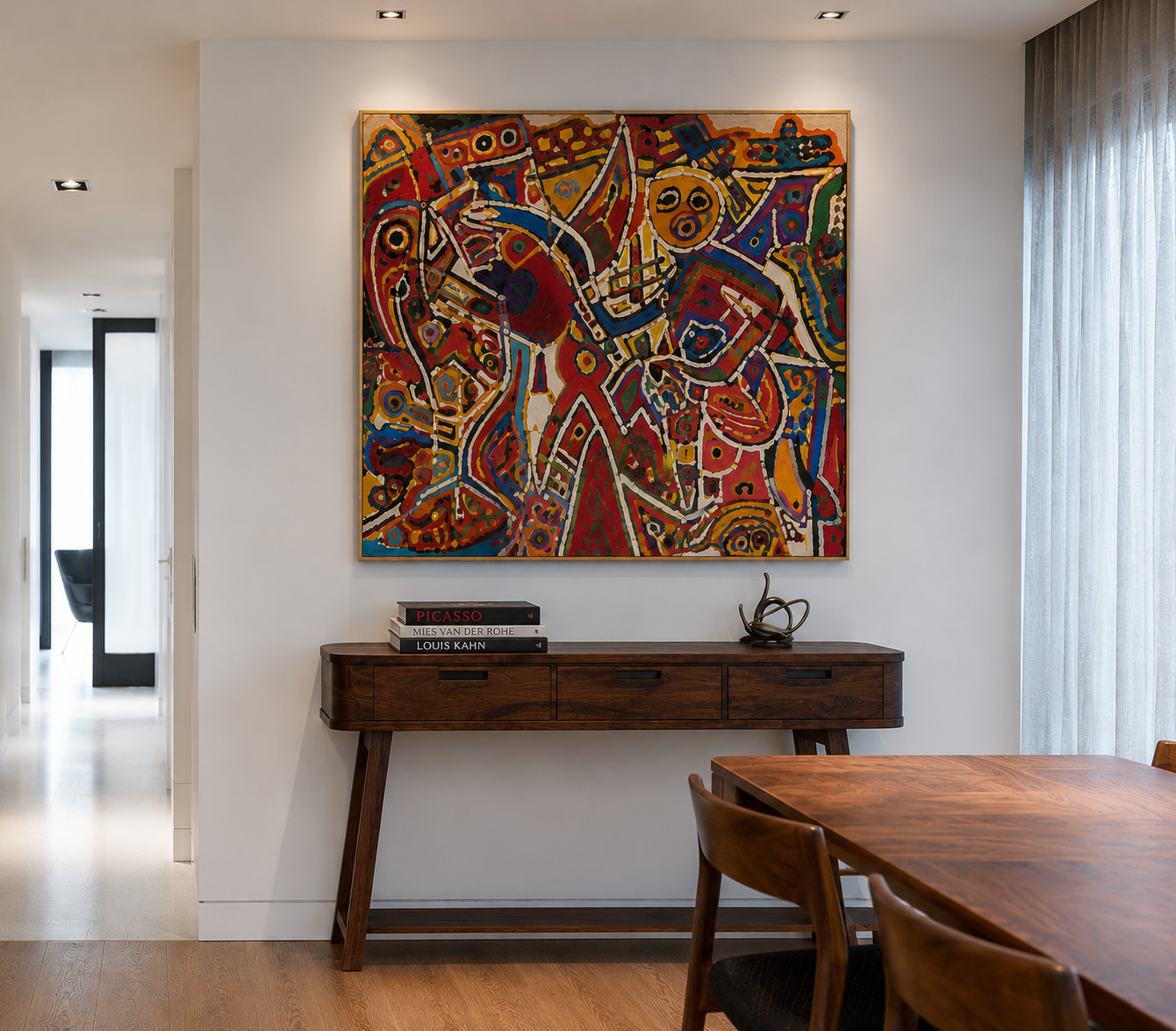 A modern living room corner with a colorful abstract painting hanging on the wall above a wooden sideboard. The sideboard has a sculpture on it and a stack of art books. To the right, sheer curtains cover a window, and part of a wooden dining table w