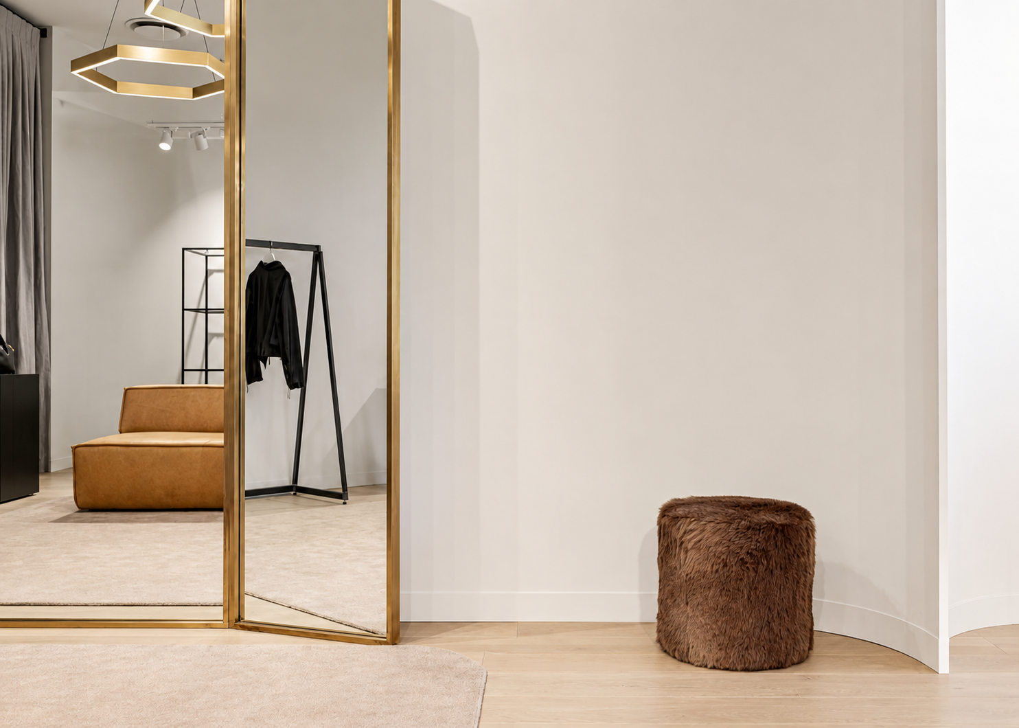 Modern interior with a gold-framed mirror, a brown furry ottoman, a tan seating area, and a clothing rack with black clothes in the background.