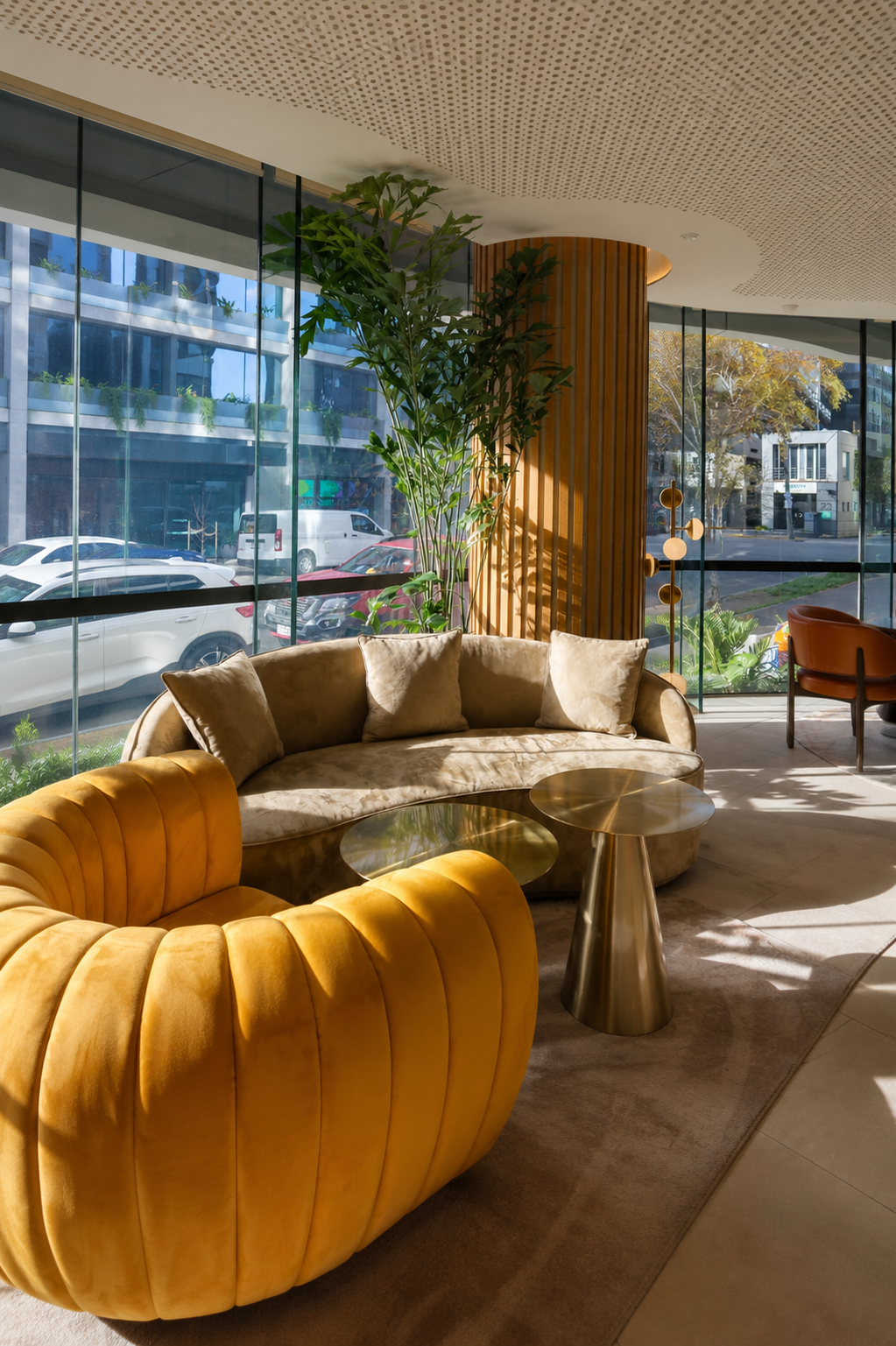 Modern lobby with large windows, beige and mustard velvet couches, gold side tables, plants, and a decorative wood-and-metal wall.