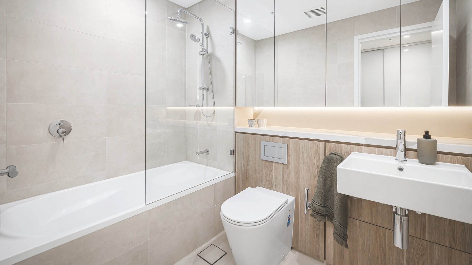 Modern bathroom with a bathtub, shower, toilet, and sink, featuring beige tiles and a large mirror with built-in lighting.